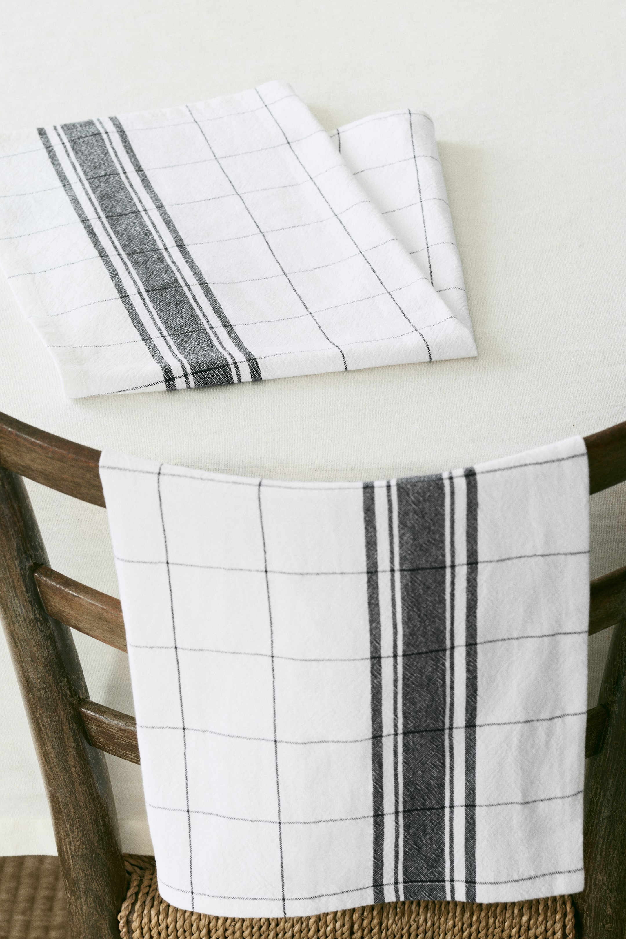View larger image: Two fabric tea towels are showcasing white woven material adorned with dark grey patterns. One towel is neatly folded, revealing a broad vertical stripe and a subtle grid pattern, while the second towel is draped, also featuring the same vertical stripe and grid patterns.