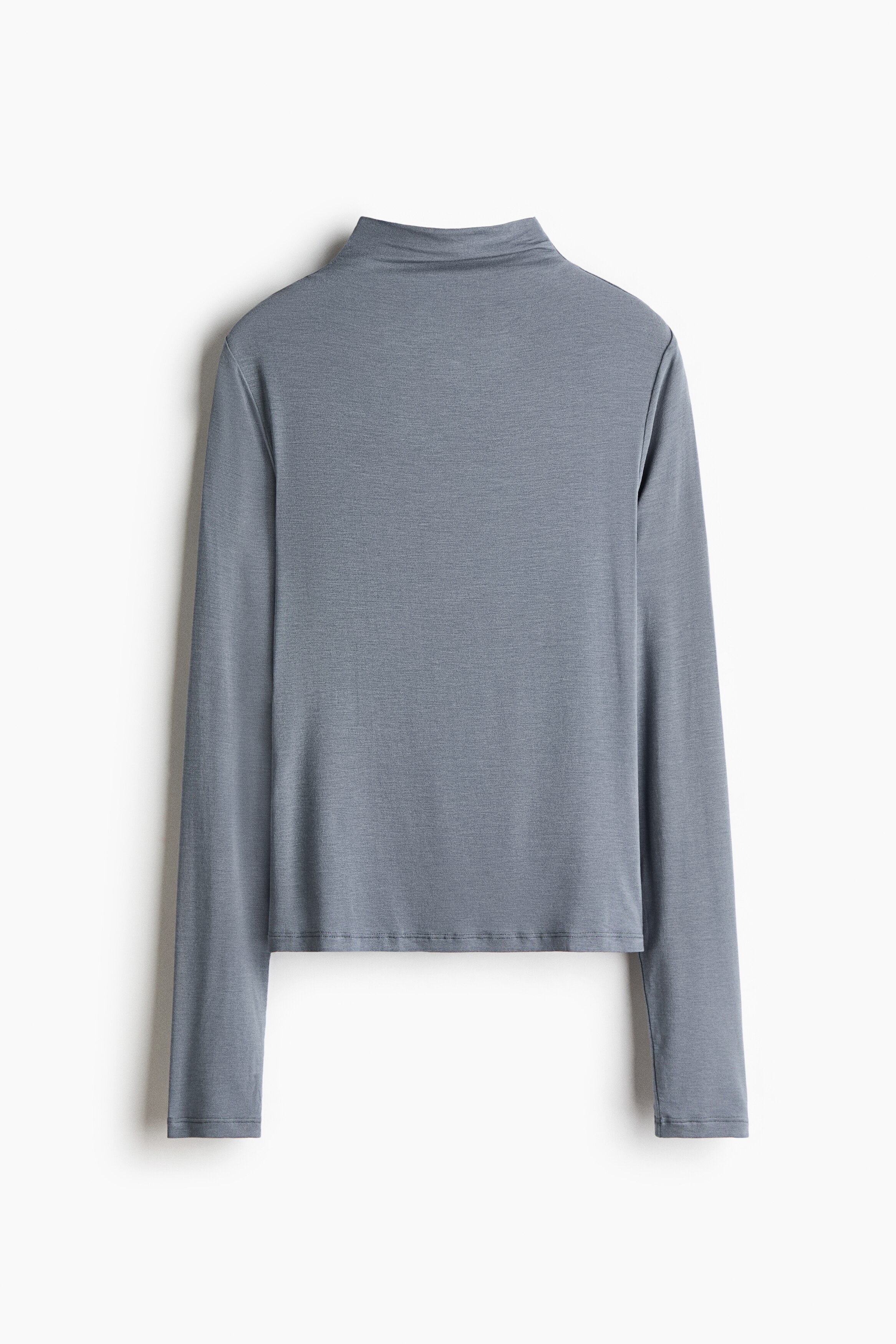 A blue-grey turtleneck base layer top with extra-long sleeves is being worn by the model, who is standing in profile and looking down. The model is resting one hand near the waistband of a pair of light grey sweatpants, highlighting the top's straight-cut hem.