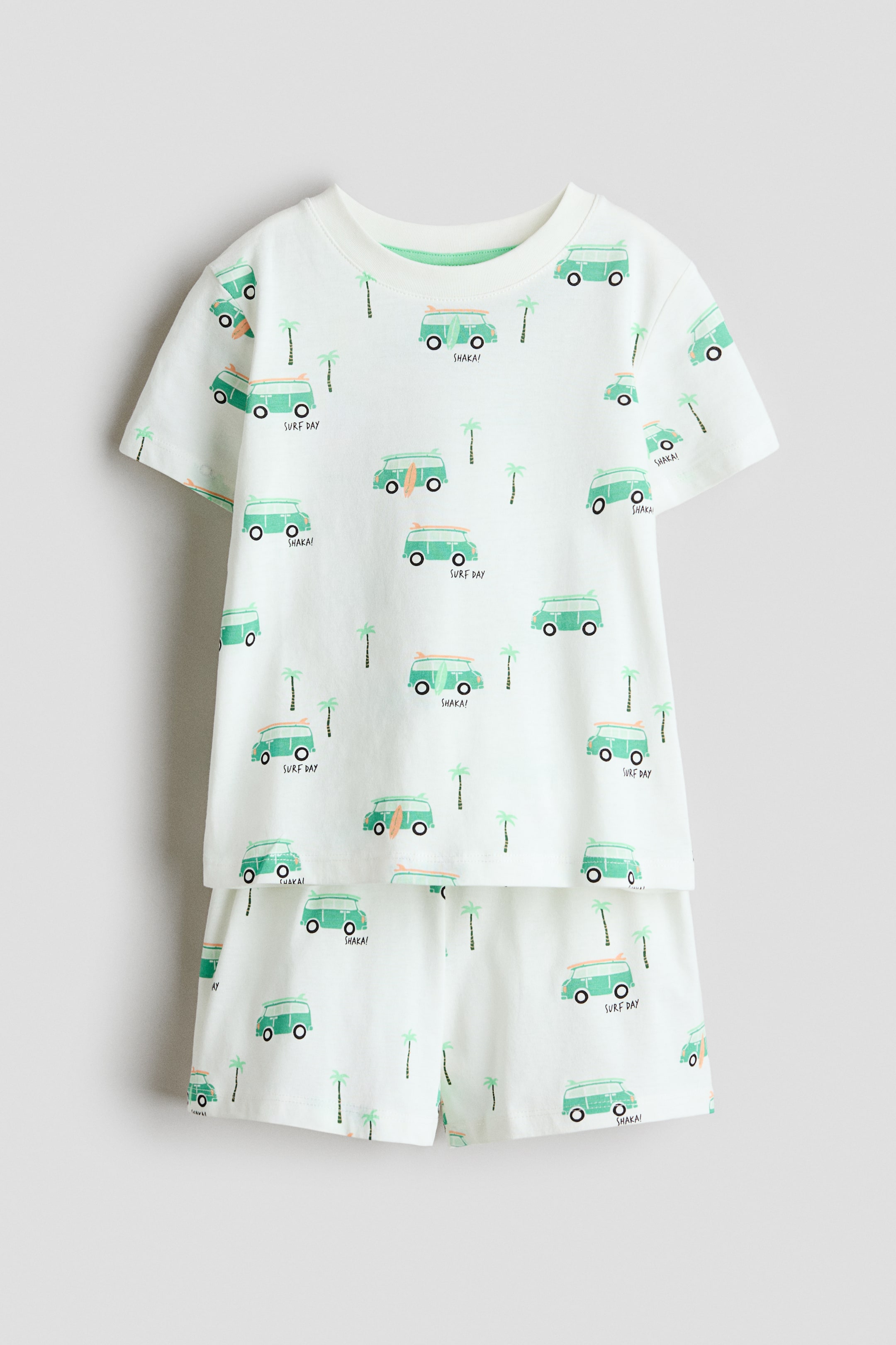 View larger image: Front-facing white pyjama set featuring a short-sleeved T-shirt and matching shorts, both adorned with a repeating pattern of light green surf vans, some with surfboards, small palm trees, and text like 'SURF DAY' and 'SHAKA!'. The T-shirt is accentuated with a white ribbed trim around its round neckline.