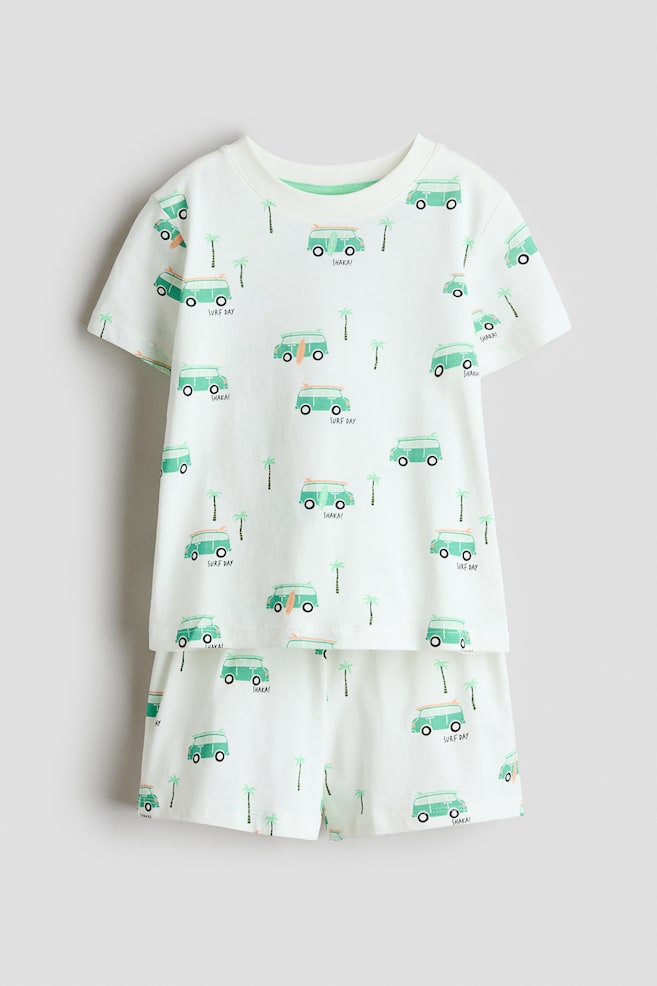 A front-facing white pyjama set features a short-sleeved T-shirt and matching shorts, both adorned with a repeating pattern of light green surf vans, some with surfboards, small palm trees, and text such as 'SURF DAY' and 'SHAKA!'. The T-shirt showcases a white ribbed trim around its round neckline. - 1