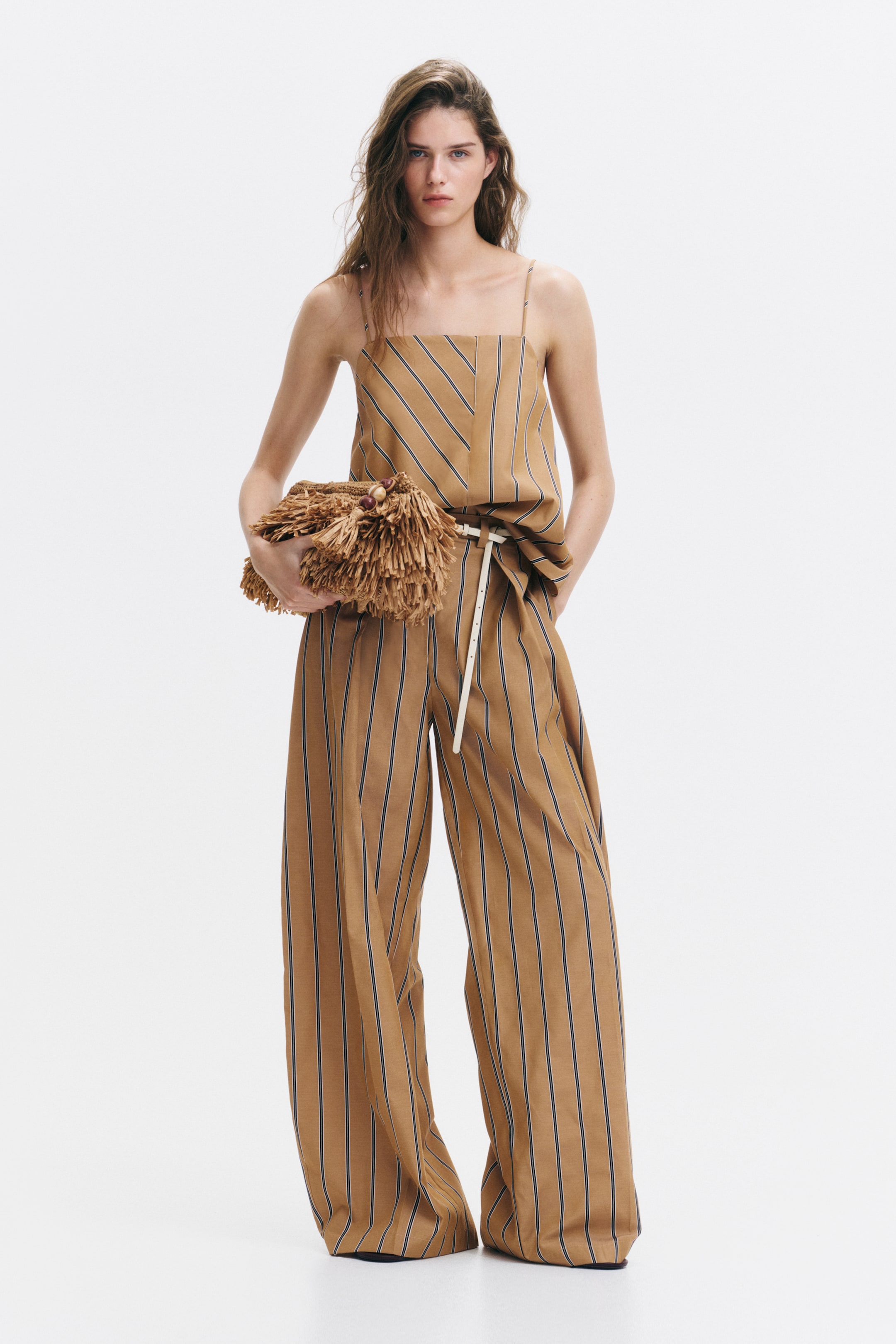 Visualizza l'immagine ingrandita: Pantaloni a vestibilità ampia in tessuto intrecciato color tan con righe verticali scure, indossati dalla modella, caratterizzati da gambe a palloncino con pieghe sul davanti e tasche laterali discrete. La mano sinistra della modella è appoggiata in una delle tasche laterali, e indossa anche un top a righe abbinato e una sottile cintura bianca.