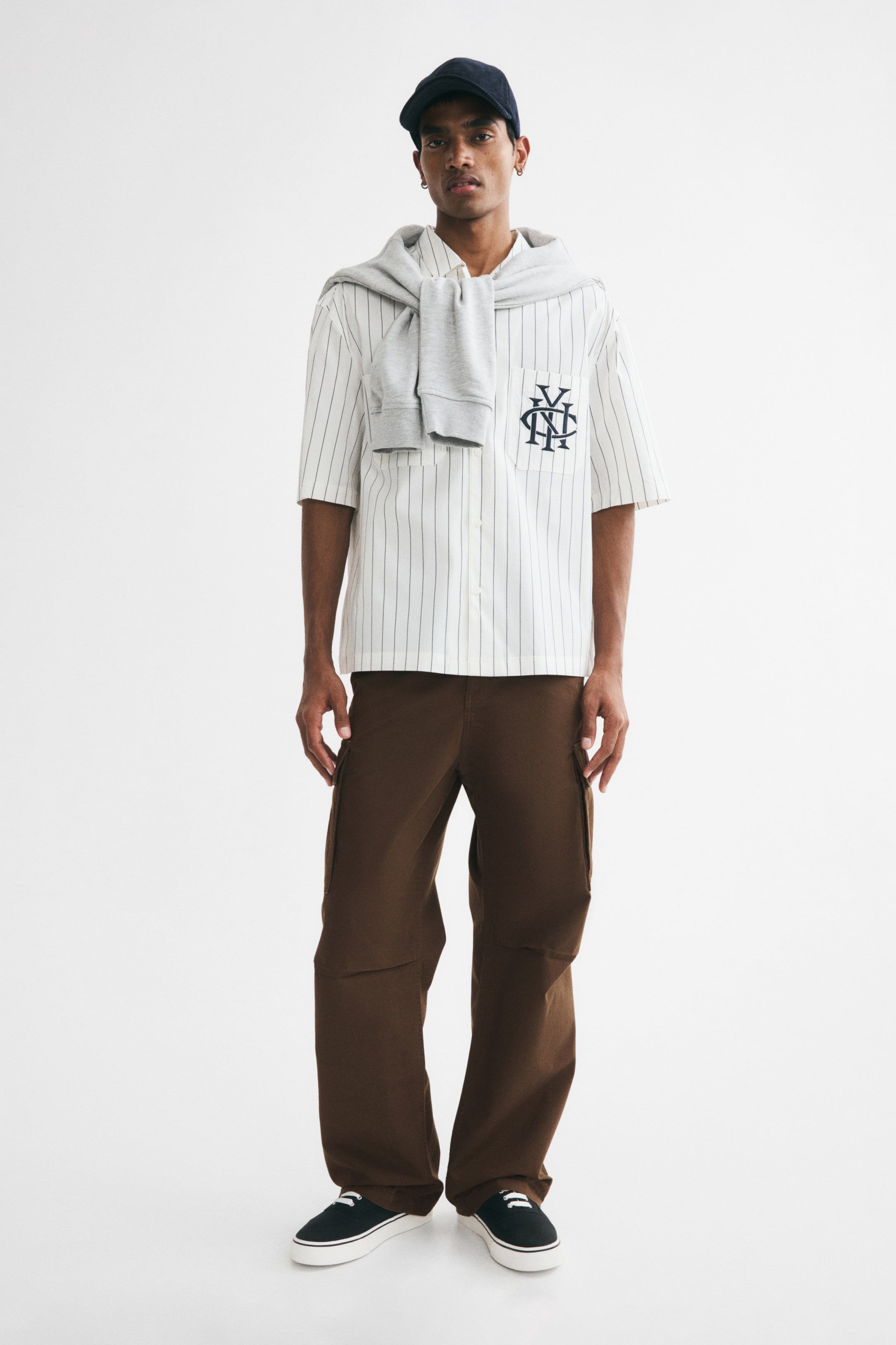 View larger image: The model is wearing brown wide-leg trousers, which feature diagonal side pockets, flapped cargo pockets on the thighs, and darts at the knees. These trousers offer a relaxed fit and are styled with black trainers.