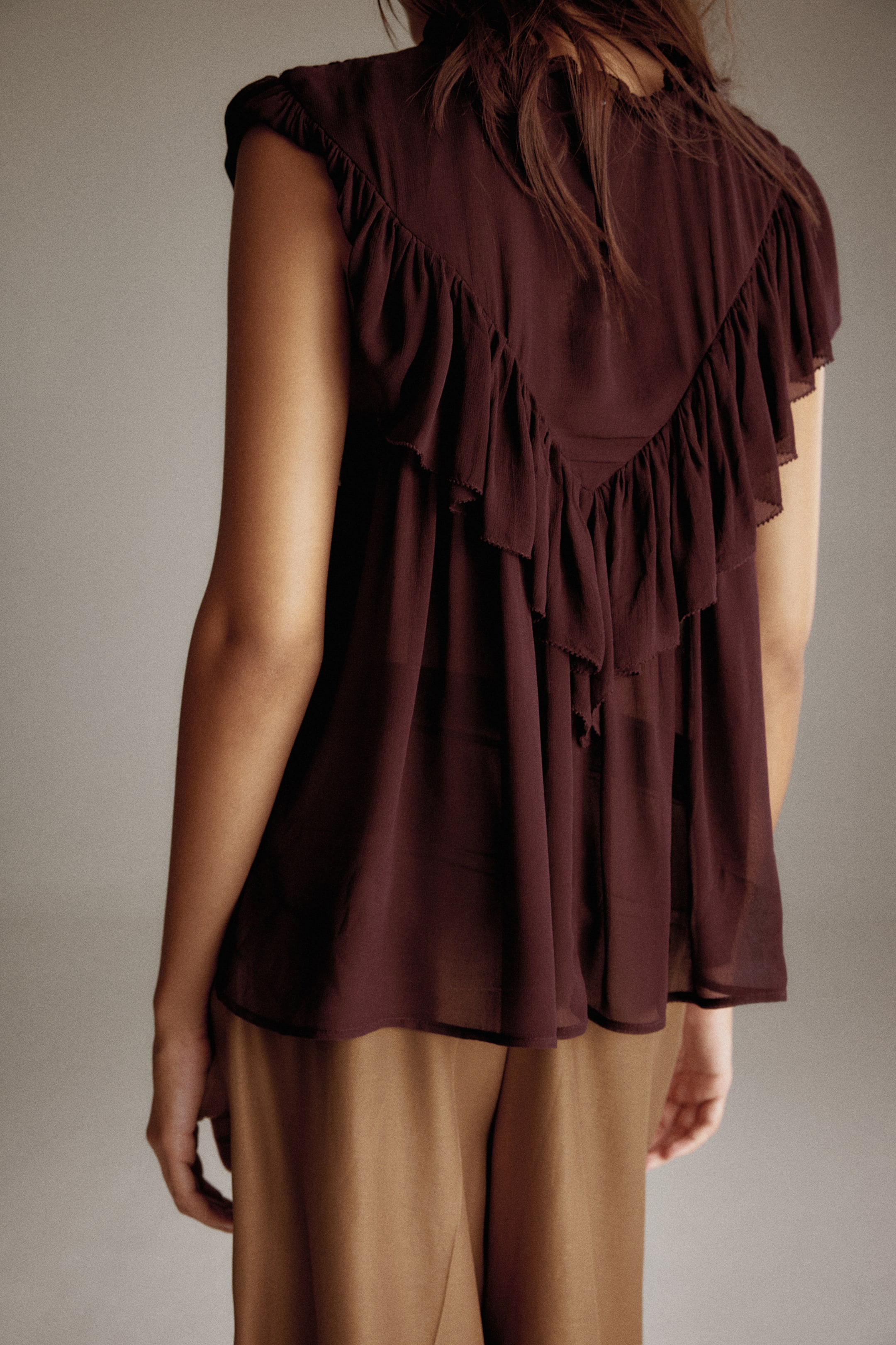 View larger image: A deep reddish-brown sleeveless top is being shown from the back, featuring multiple layered ruffles forming a V-shape. The sheer fabric is flowing loosely, with the ruffles adding volume to the top.
