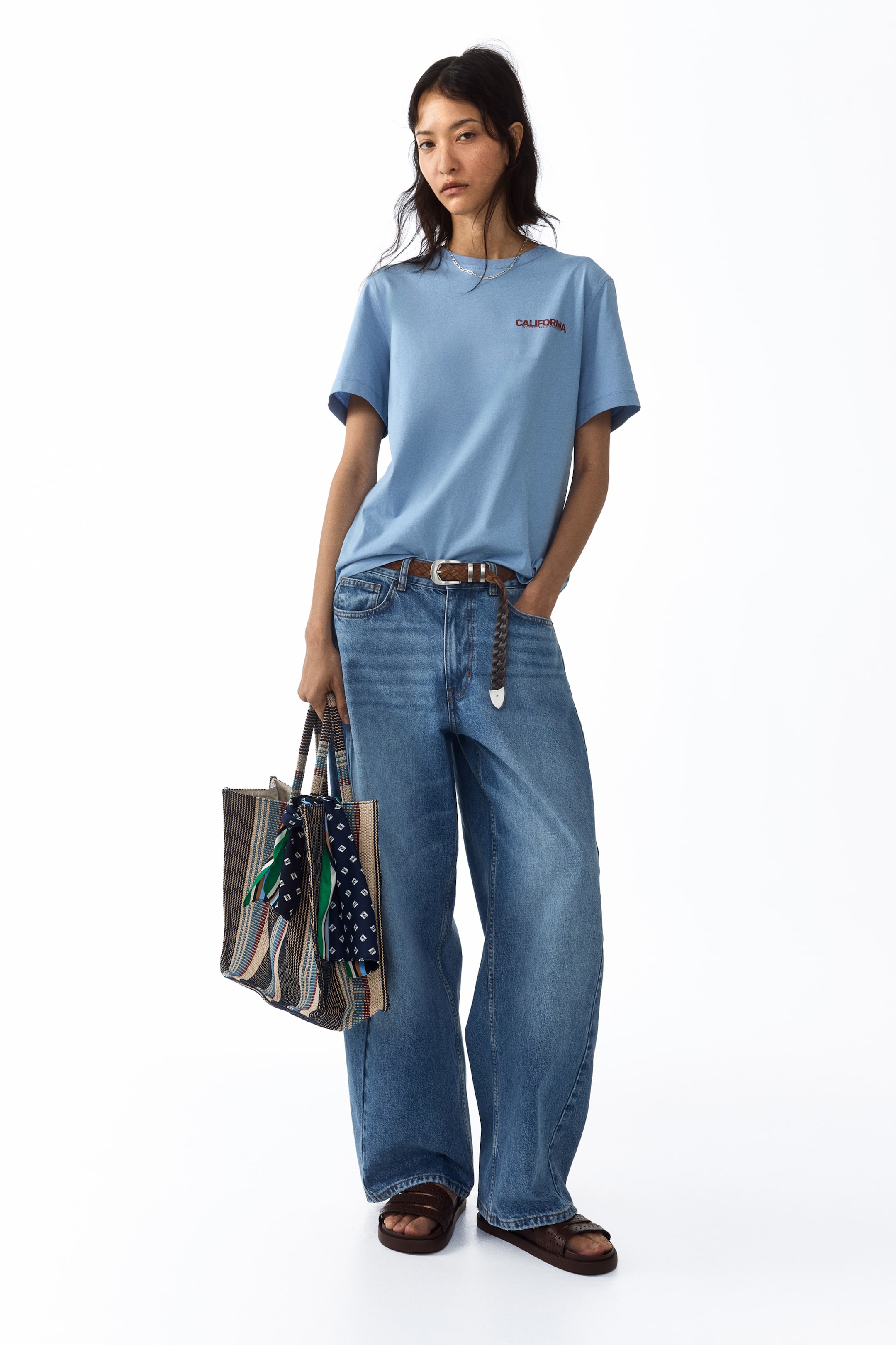 Voir une image plus grande: Un T-shirt classique bleu clair en jersey de coton, arborant un motif imprimé "CALIFORNIA" rouge sur la poitrine, avec un col rond et un ourlet droit, est porté par le mannequin. Le T-shirt est rentré dans un jean à jambes larges et stylé avec une ceinture tressée marron.