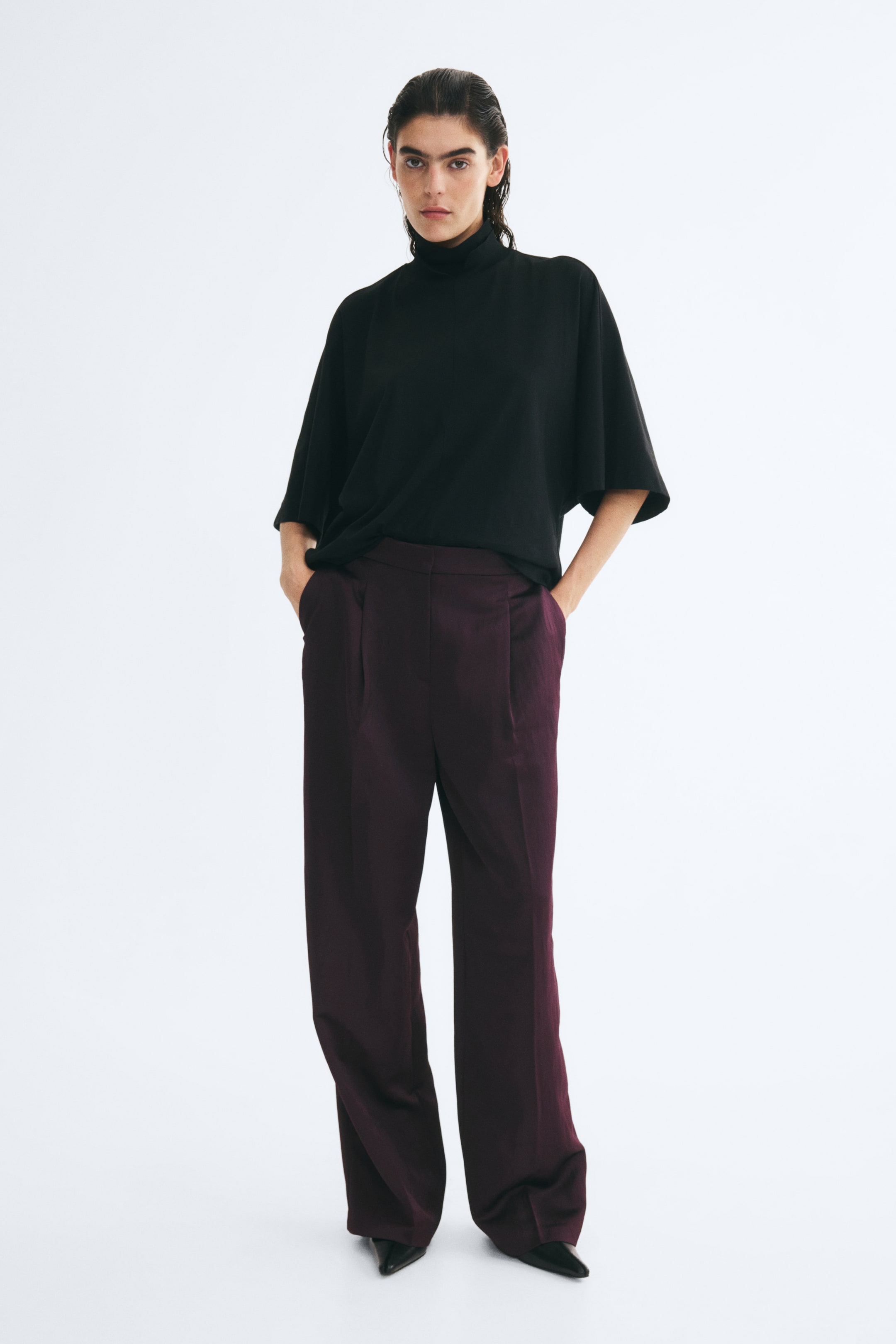 View larger image: Wide-leg, high-waisted pants in dark purple, lightly crinkled fabric are worn by the model, who stands with hands resting in the diagonal side pockets. The loose-fit trousers feature front pleats and pressed creases, styled with a black mock-neck top and black pointed-toe shoes.