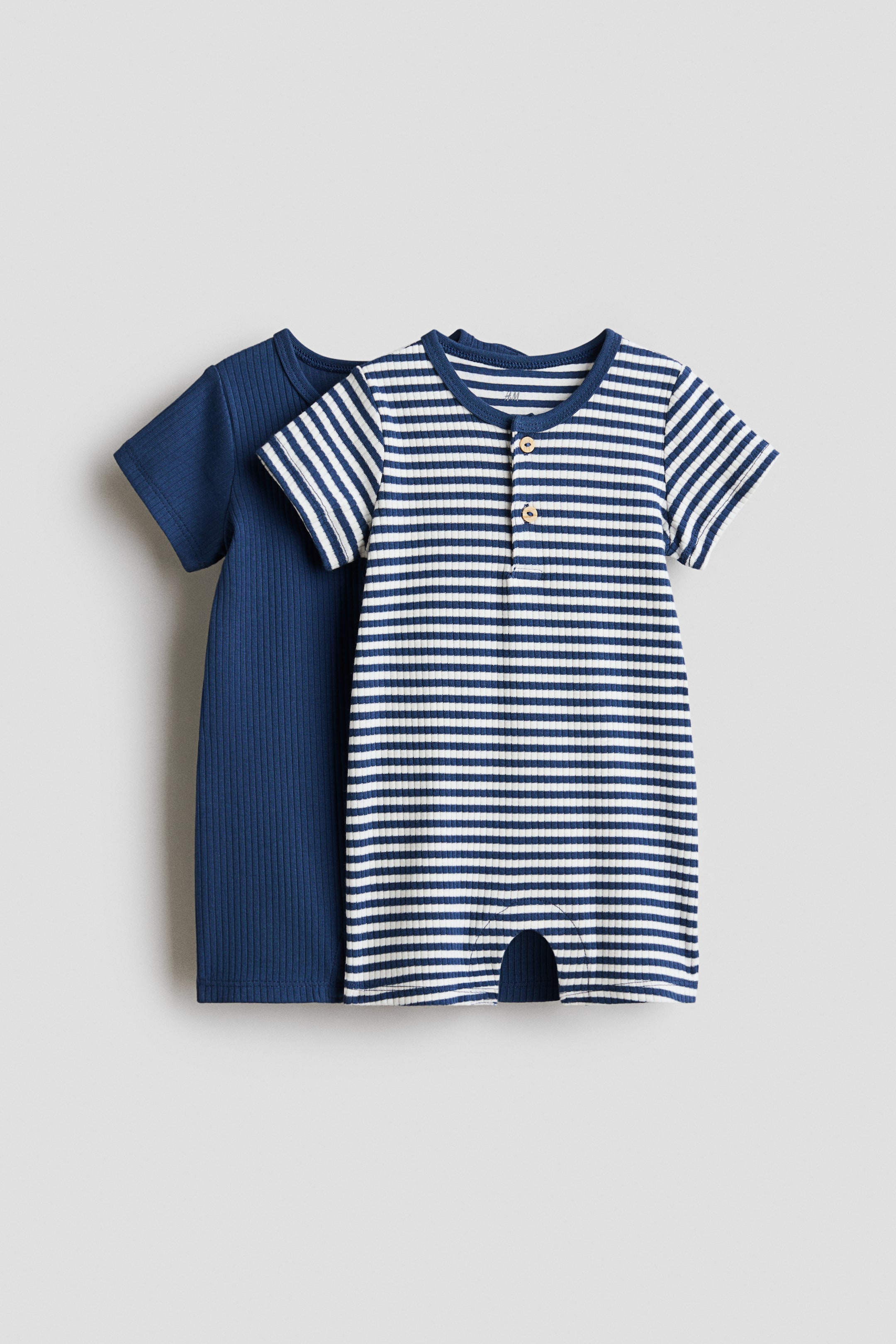 View larger image: Two ribbed baby rompers are on display. The front romper showcases a white and navy blue horizontal stripe pattern, complemented by short sleeves, a round neckline, a partial button placket with two buttons, and a crotch cutout. Behind it, a solid navy blue romper with a ribbed texture and short sleeves adds a layered look.