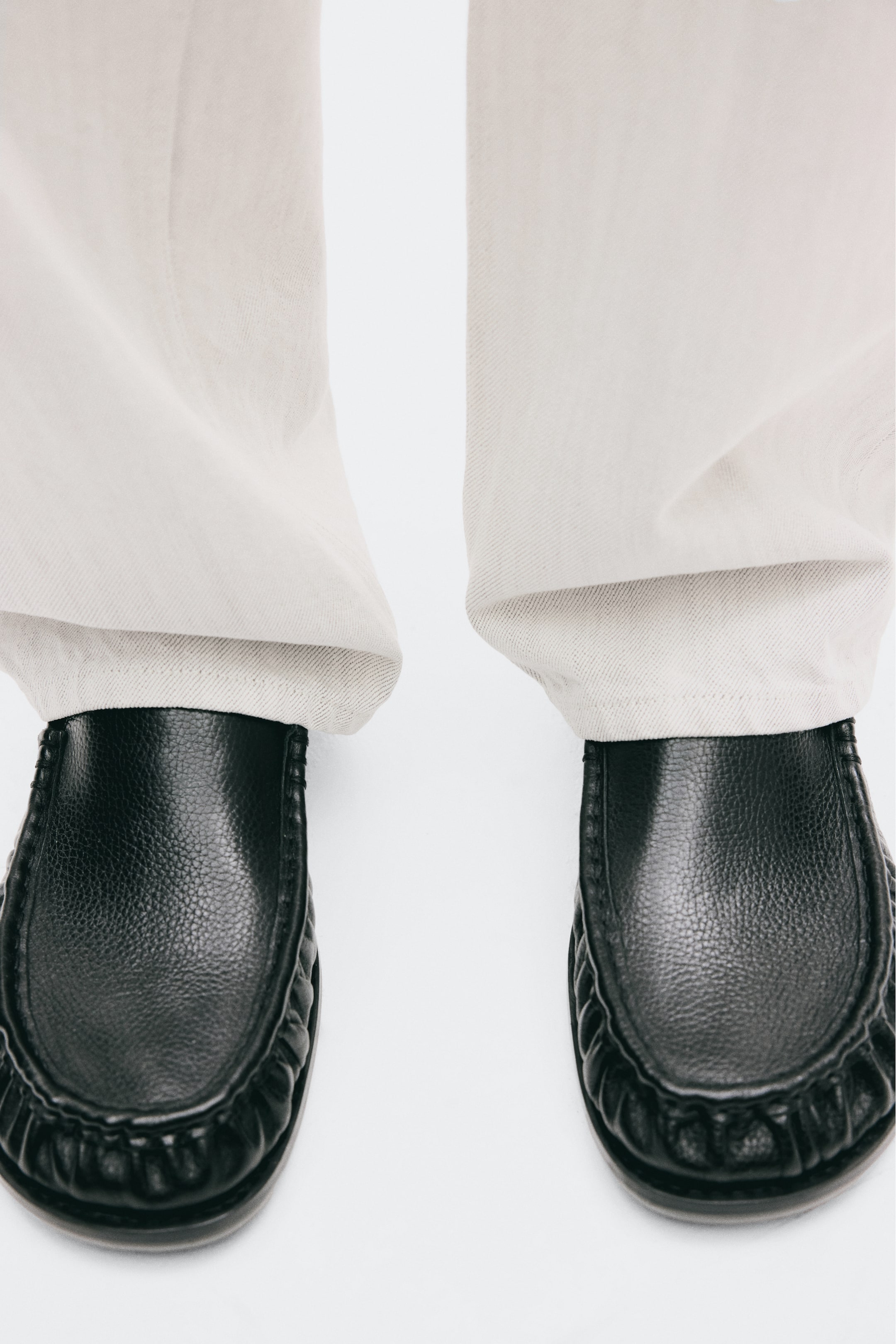 Ver imagen más grande: Unos mocasines de piel negra, con costura tipo mocasín y frunces en la parte delantera, los lleva puestos la modelo. Unos pantalones de color claro descansan por encima de los mocasines.