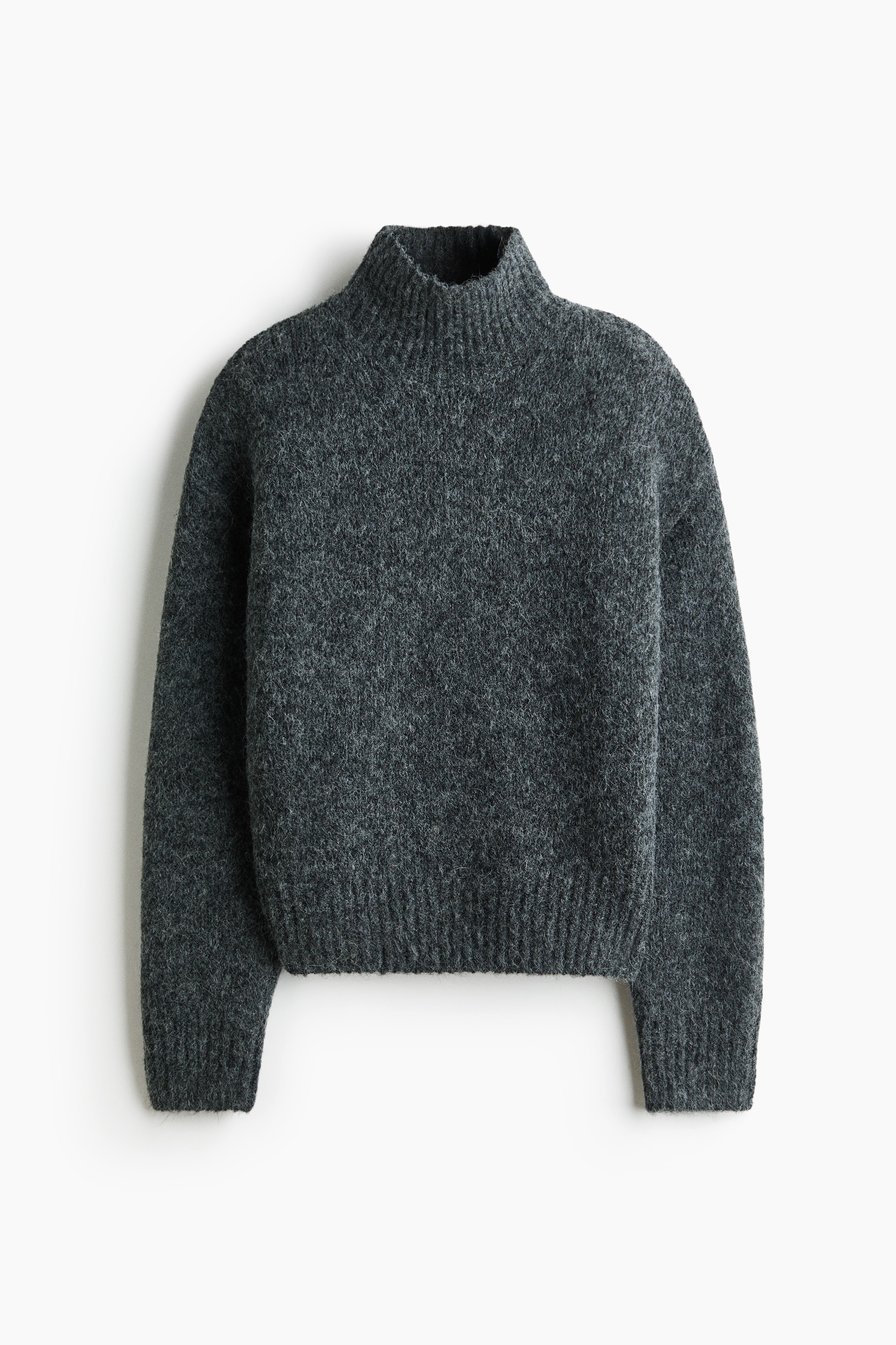 The model is wearing a dark grey sweater in a soft, brushed knit, featuring a high ribbed neck and dropped shoulders. The oversized sweater is partially tucked into wide-leg white trousers at the front.