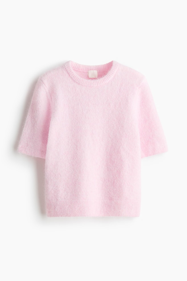 A pink fluffy sweater is laid flat, displaying a crew neck, short sleeves with ribbed cuffs, and a ribbed hem. A light-colored tag is visible at the back of the neck. - 1