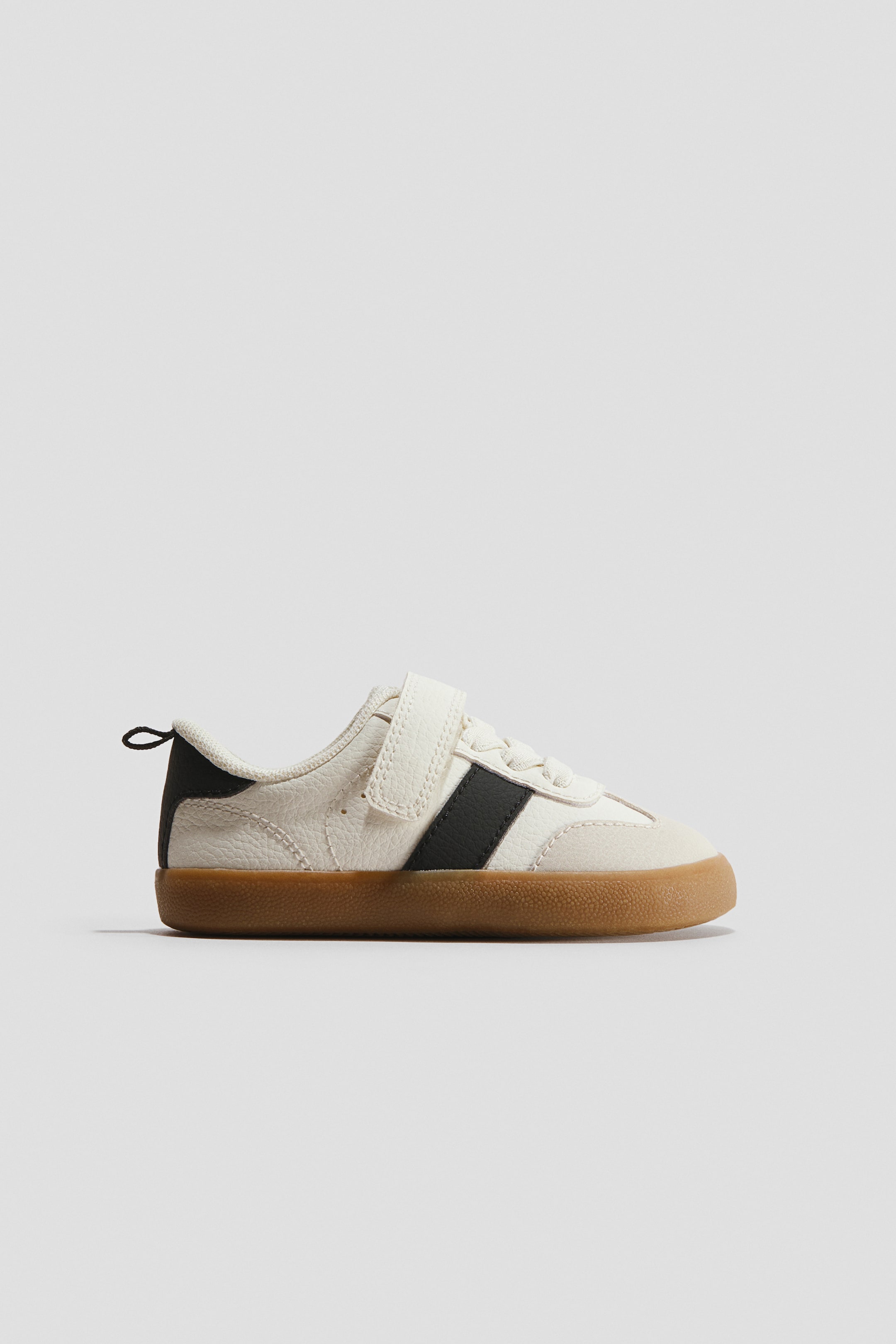 View larger image: A barefoot trainer is facing right in a side profile view, displaying an off-white textured leather upper with a light beige suede toe cap and a contrasting black side stripe. The shoe features a single hook-and-loop strap closure over elastic laces and rests on a flat, textured brown gum sole.
