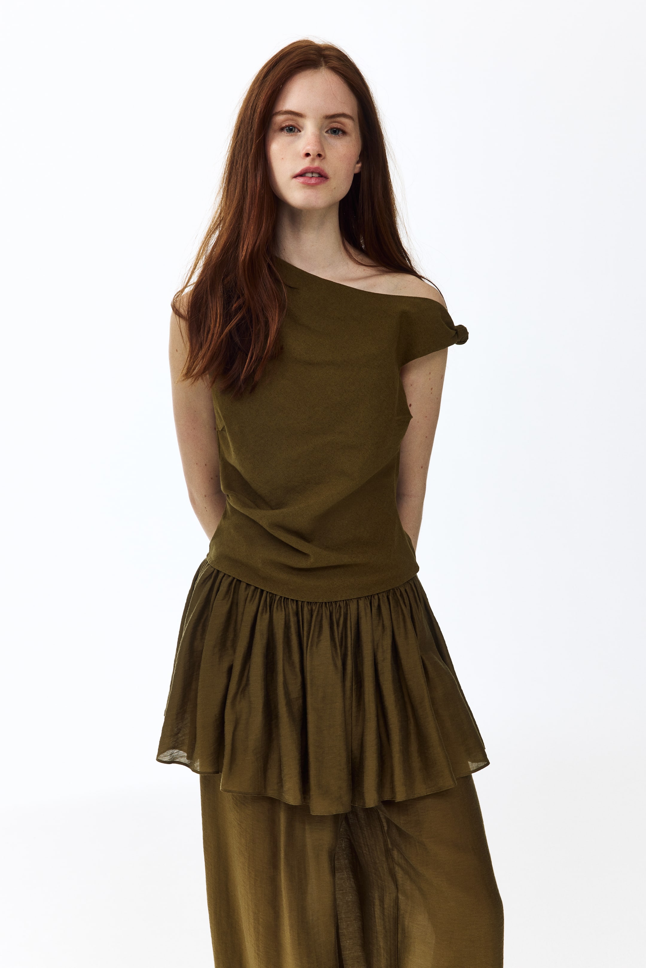 View larger image: Olive green asymmetrical top yang menghadap ke depan, menampilkan rekaan off-the-shoulder di sebelah kanan dengan perincian simpul, dipadankan dengan bahagian bawah berlapis warna hijau zaitun yang sepadan. Bahagian bawah ini menonjolkan lapisan gaya peplum yang berkumpul dan lapisan panjang yang mengalir di bawah dengan belahan yang kelihatan.