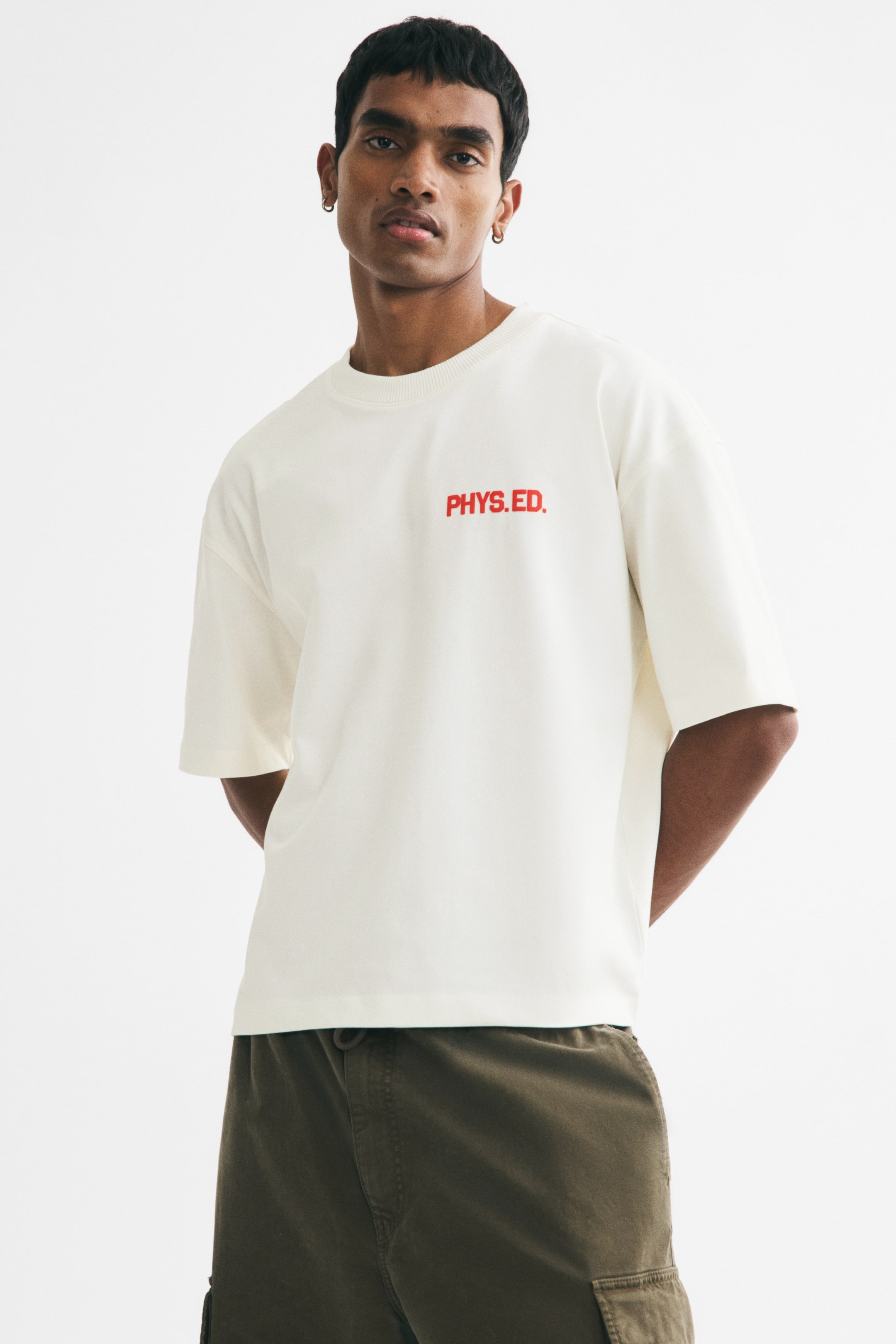 View larger image: An off-white boxy-style T-shirt with an oversized, baggy fit, a round, rib-trimmed neckline, dropped shoulders, and a red 'PHYS.ED.' print on the chest is being worn. The model is standing with hands behind the back.