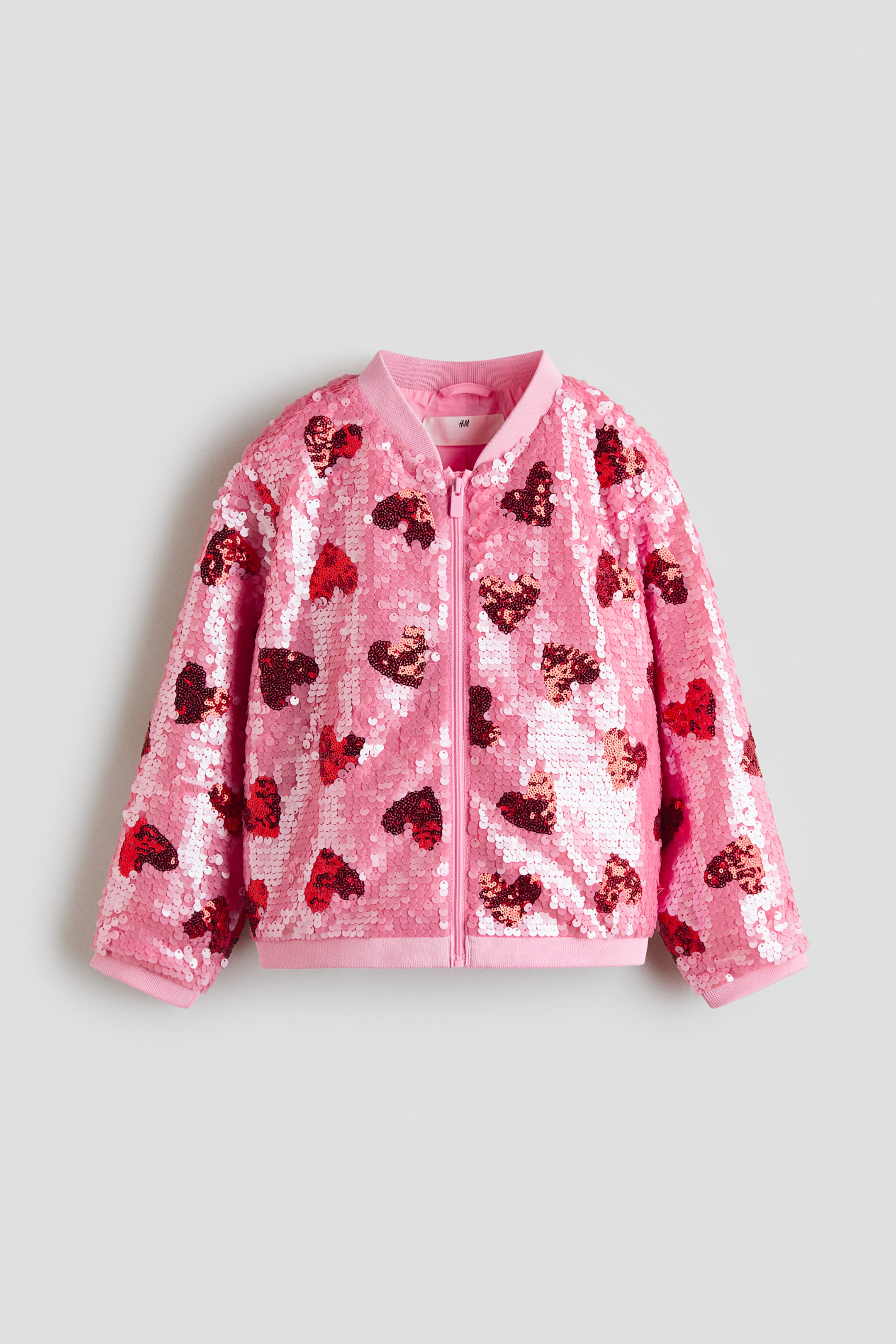 Größeres Bild anzeigen: Eine rosa Bomberjacke zeigt ihre Vorderseite, gefertigt aus Pailletten-Mesh mit durchgehenden Herzmustern in verschiedenen Rosa- und Rottönen. Sie verfügt über einen niedrigen, gerippten Kragen, einen Frontreißverschluss, lange Ärmel mit schmalen, gerippten Abschlüssen an den Bündchen und am Saum und ist gefüttert.