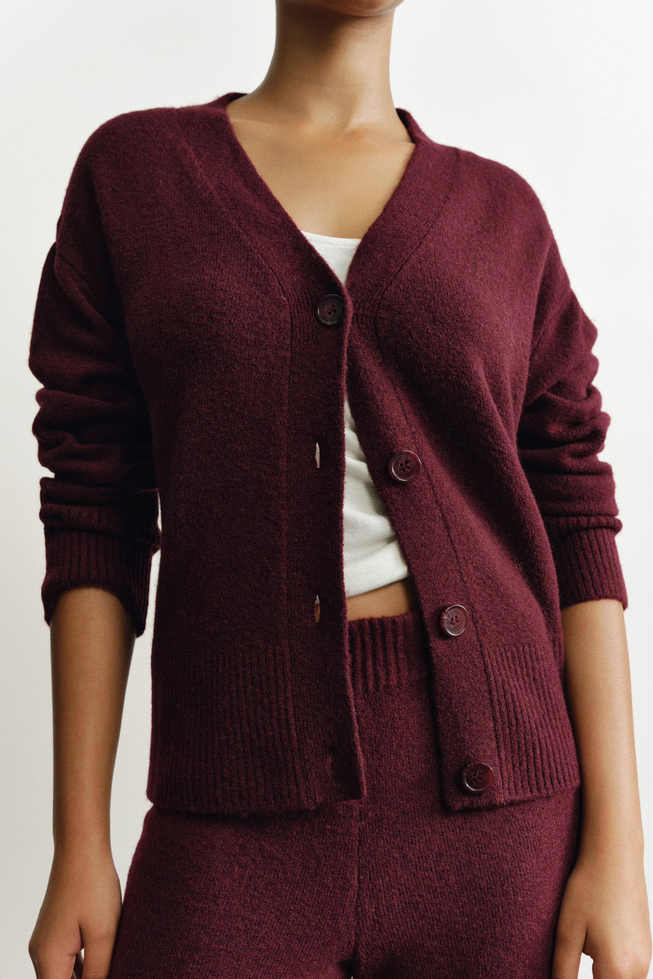 View larger image: A burgundy knit cardigan featuring a V-neck, dropped shoulders, and a button-down front is worn by the model over a white top and matching knit bottoms. The cardigan features ribbed detailing along the neckline, button band, cuffs, and hem, and is styled partially unbuttoned.