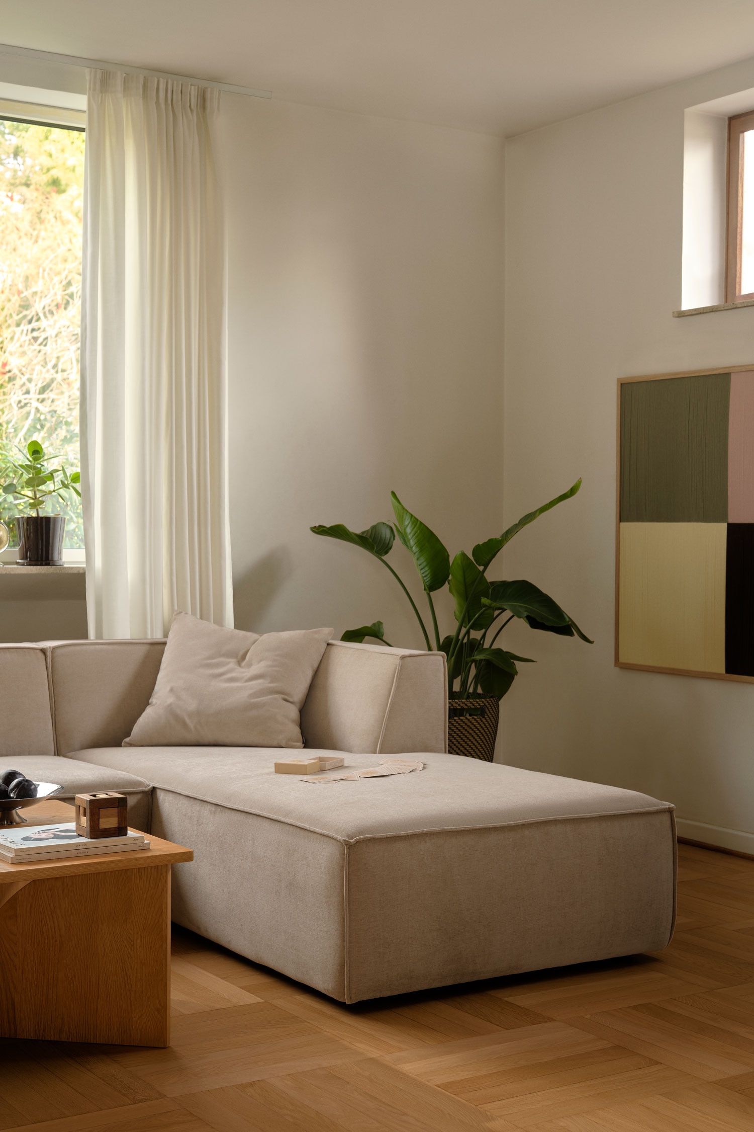 Ein modulares Ecksofa, bezogen mit hellbeigem, strukturiertem Stoff, ist frontal ausgerichtet und zeichnet sich durch ein flaches, kantiges Design aus. Das Sofa besteht aus drei modularen Elementen, darunter ein linker Eckplatz, ein mittlerer Sitz ohne Armlehnen und ein rechter Chaise-Lounge-Teil mit abgerundeten inneren RÃ¼ckenlehnen.