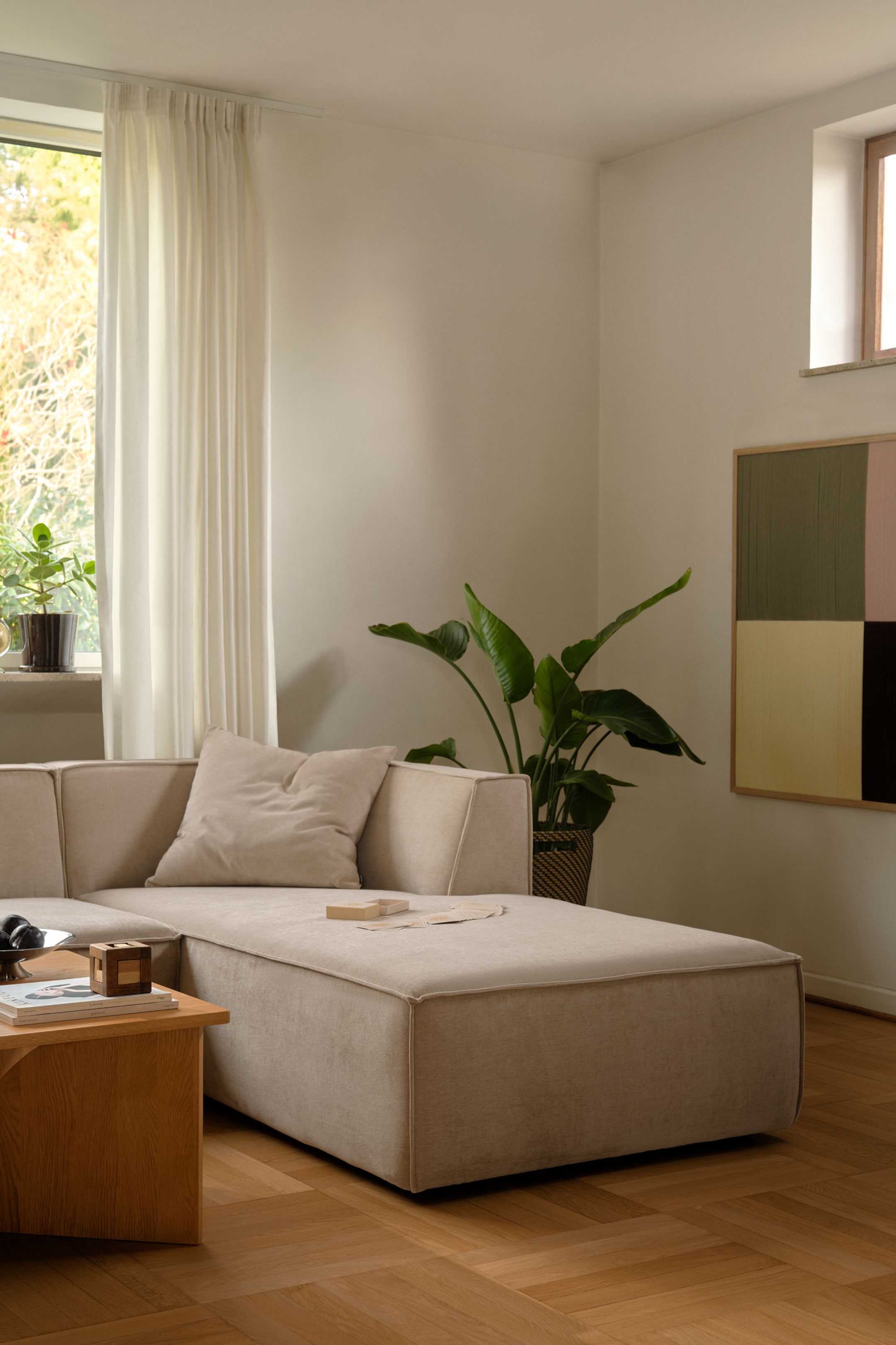 Größeres Bild ansehen: Ein modulares Ecksofa mit beigem Stoffbezug erstreckt sich zu einer langen Chaise Longue, die schräg in Richtung Vordergrund ausgerichtet ist. Das niedrig profilierte Sofa präsentiert klarlinige, kantige Polster mit Kedernähten sowie ein passendes quadratisches Zierkissen, das an der schrägen Rückenlehne anliegt.