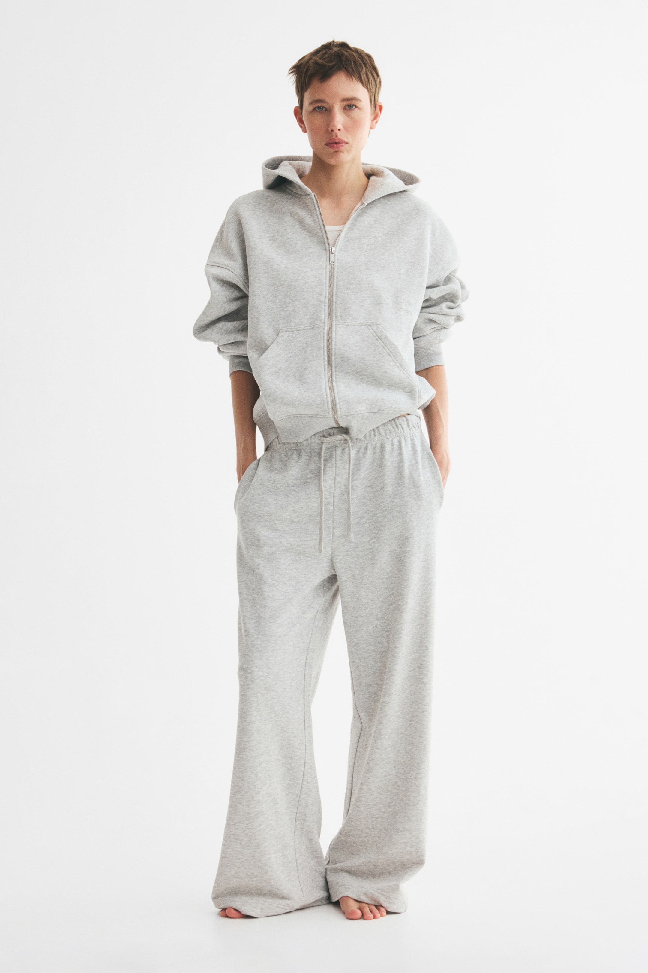View larger image: The model is wearing a light grey zip-up hoodie, featuring a relaxed fit, lined hood, dropped shoulders, and open front pockets. The hoodie is styled with matching wide-leg sweatpants.