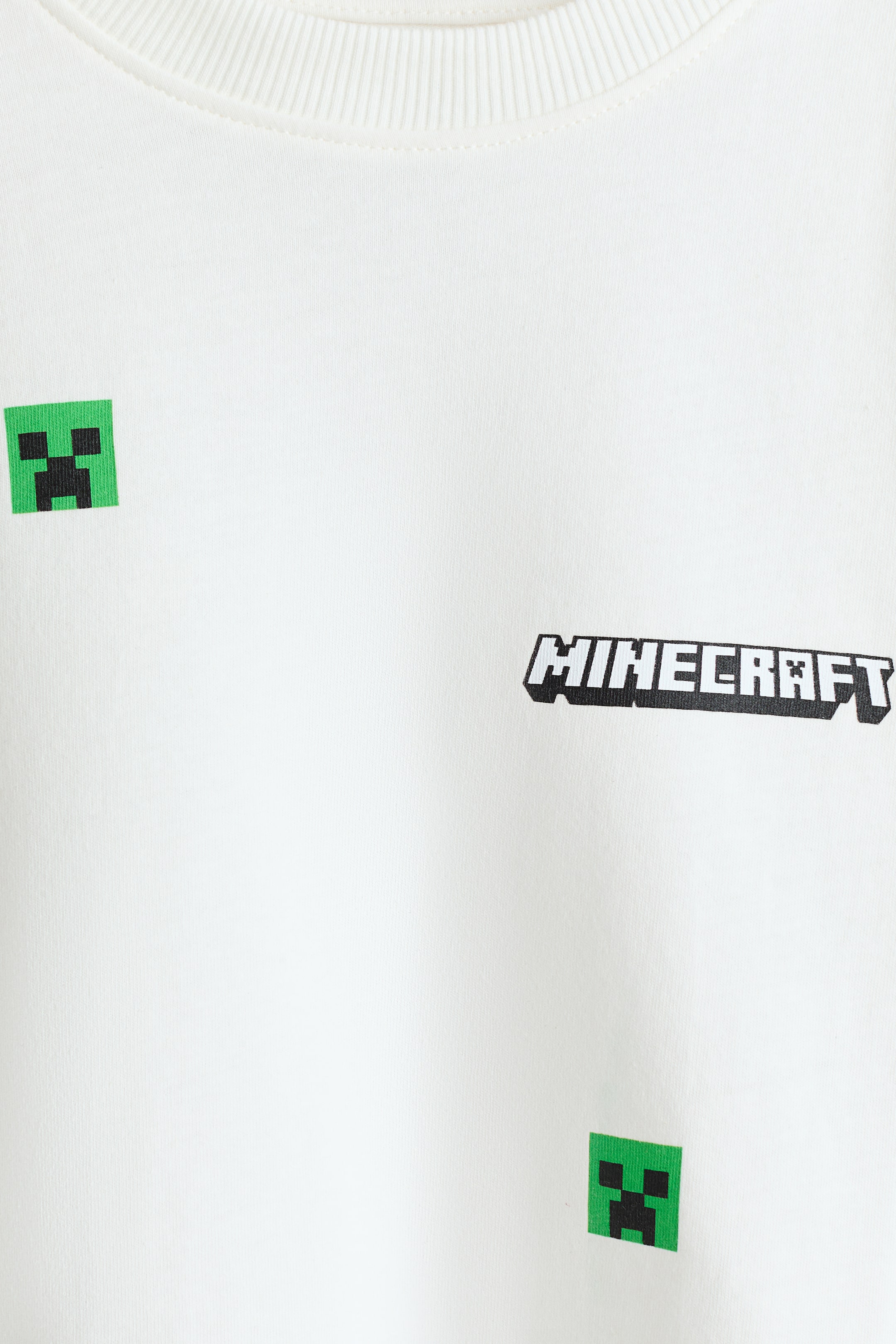 Ver imagen más grande: Una camiseta de gran tamaño en suave jersey de algodón blanco presenta un cuello acanalado. Muestra un logo negro de "MINECRAFT" y caras de Creeper pixeladas en verde dispersas en la parte delantera.