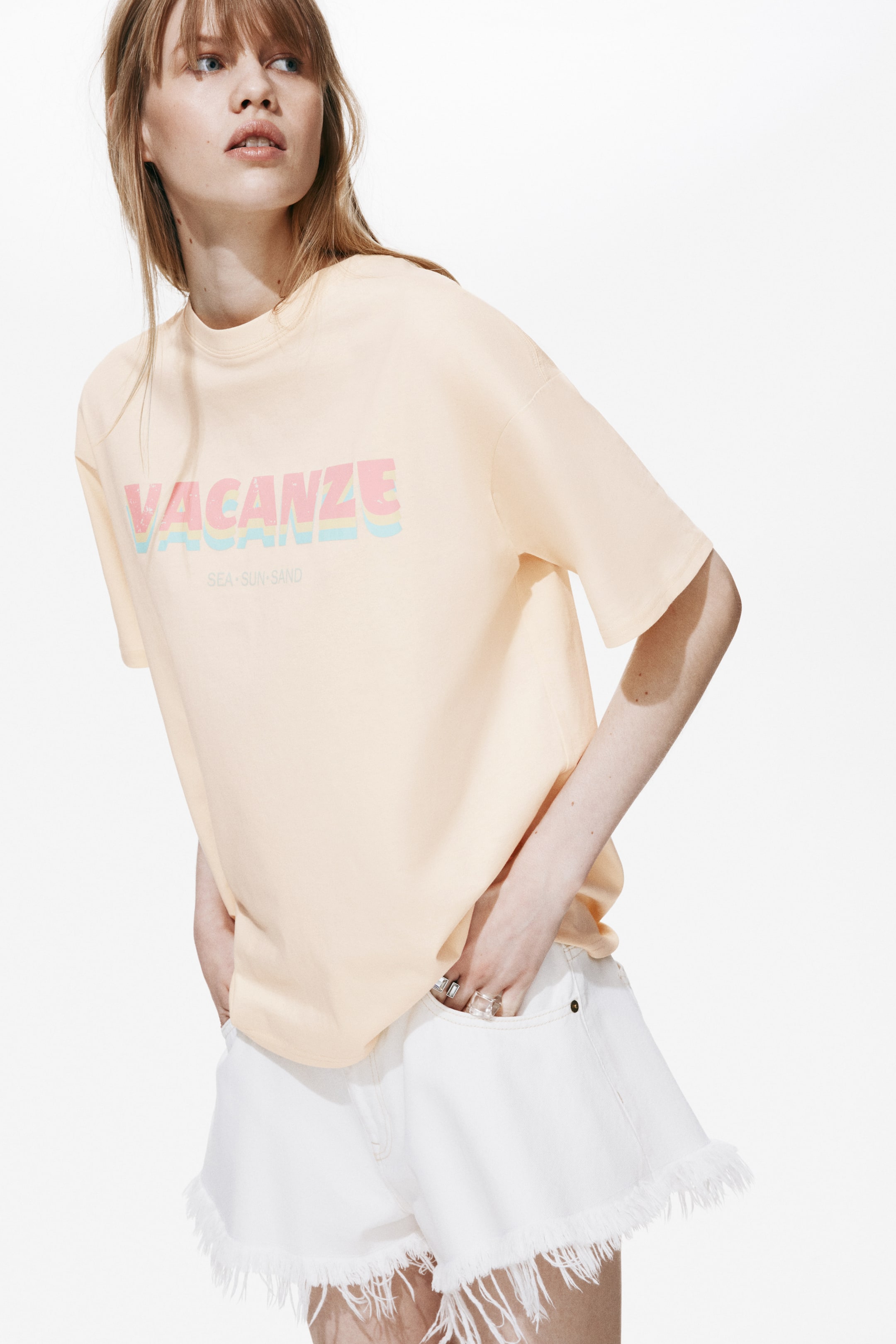 View larger image: An oversized t-shirt crafted from soft, peach-printed cotton jersey, featuring a ribbed neckline, dropped shoulders, and a straight-cut hem, is worn by the model. It showcases a vibrant "VACANZE SEA-SUN-SAND" print on the front and is partially tucked into white shorts.