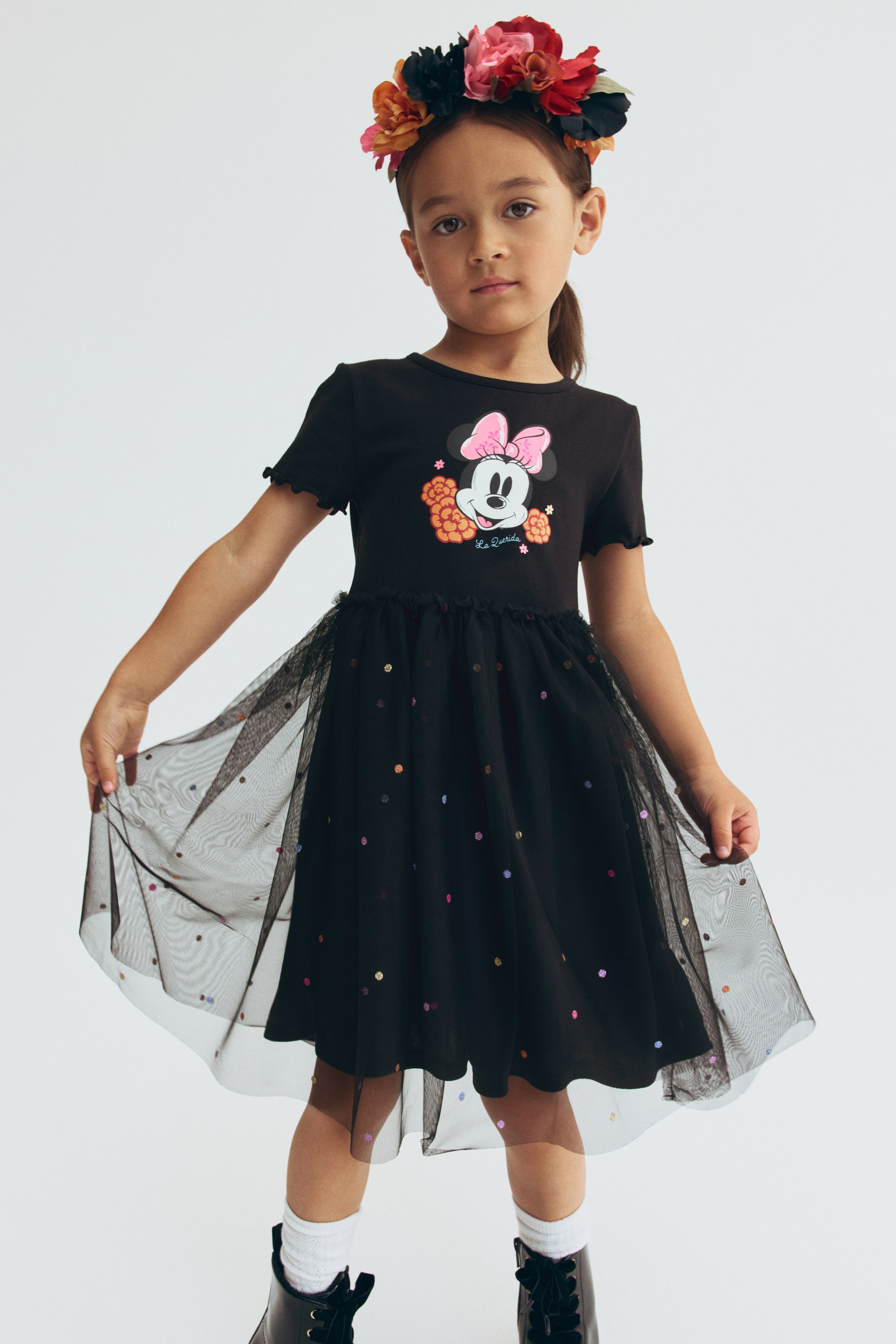 View larger image: A child is wearing a black jersey dress, shown from the front, featuring a Minnie Mouse graphic printed on the chest, a round neckline, and short sleeves with overlocked cuffs. The dress is designed with a small frill trim at the waist and a flared black tulle skirt covered in colorful, foil-printed dots.