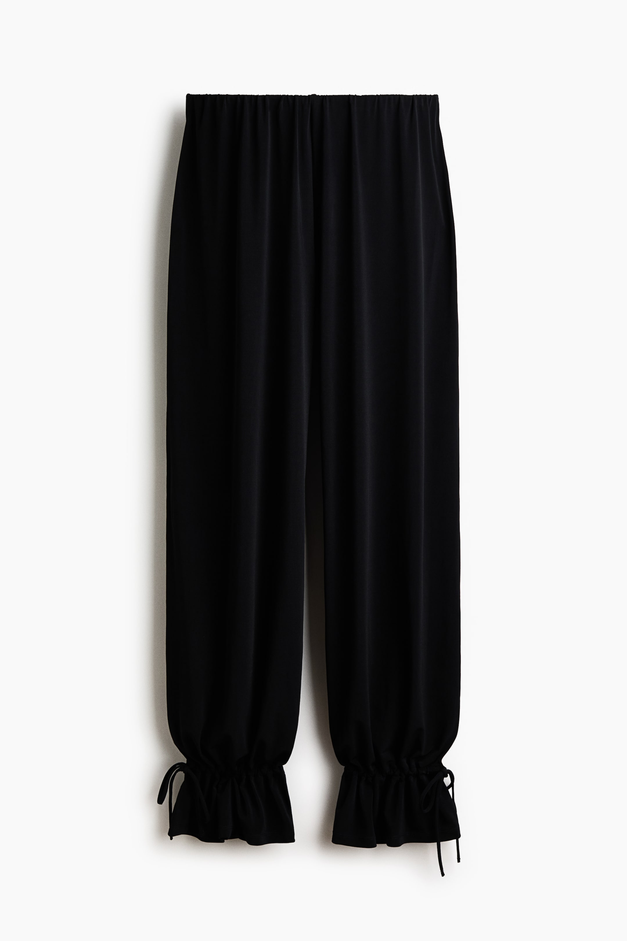 View larger image: Black trousers are showcased from a front perspective, highlighting an elasticated waistband. The wide legs cascade down to gathered ankles, which are cinched with visible ties.