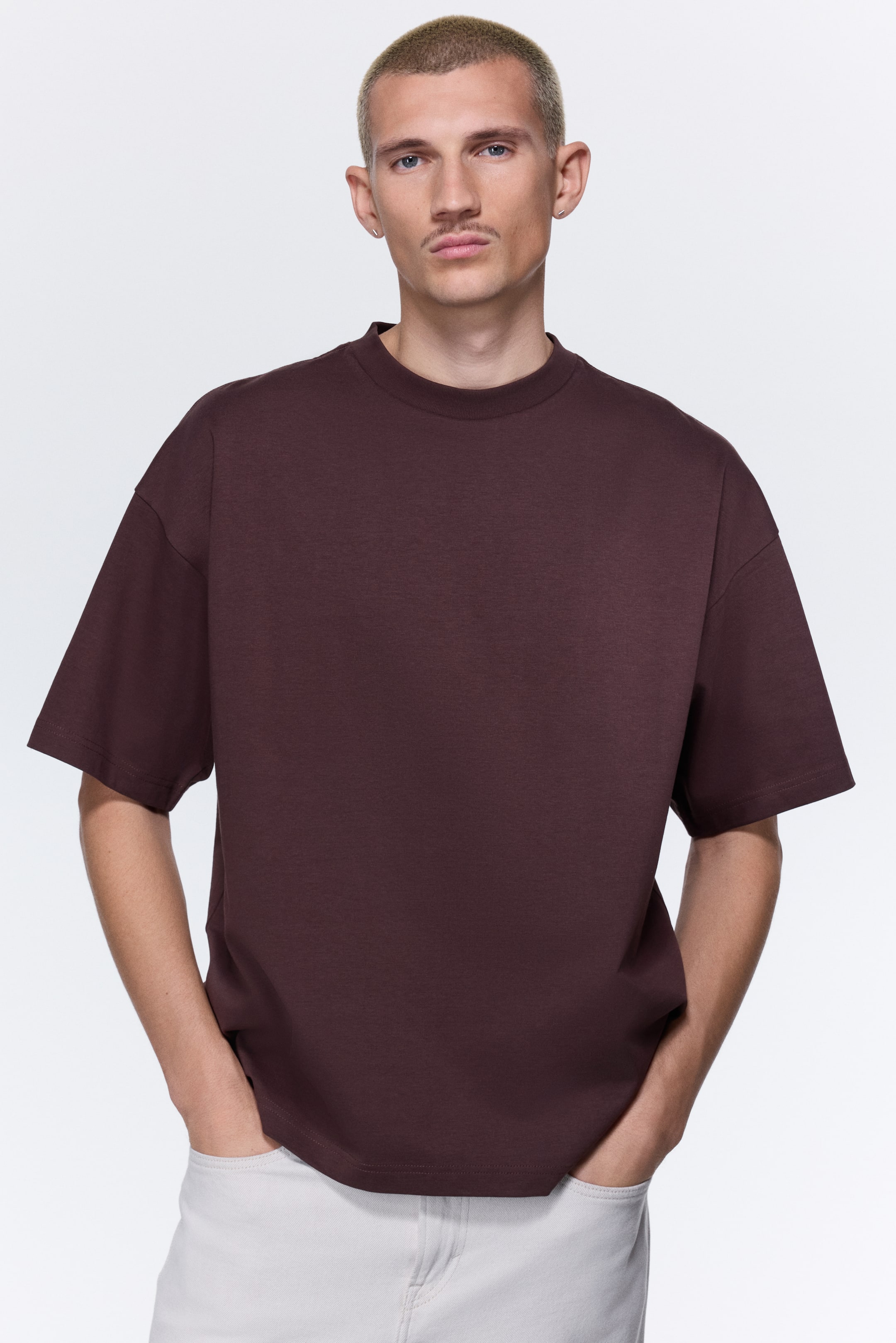 拡大画像を表示: ダークブラウンのTシャツは、ドロップショルダーと短くて幅の広い袖のオーバーサイズフィットを特徴としており、モックネックとストレートな裾がデザインされています。