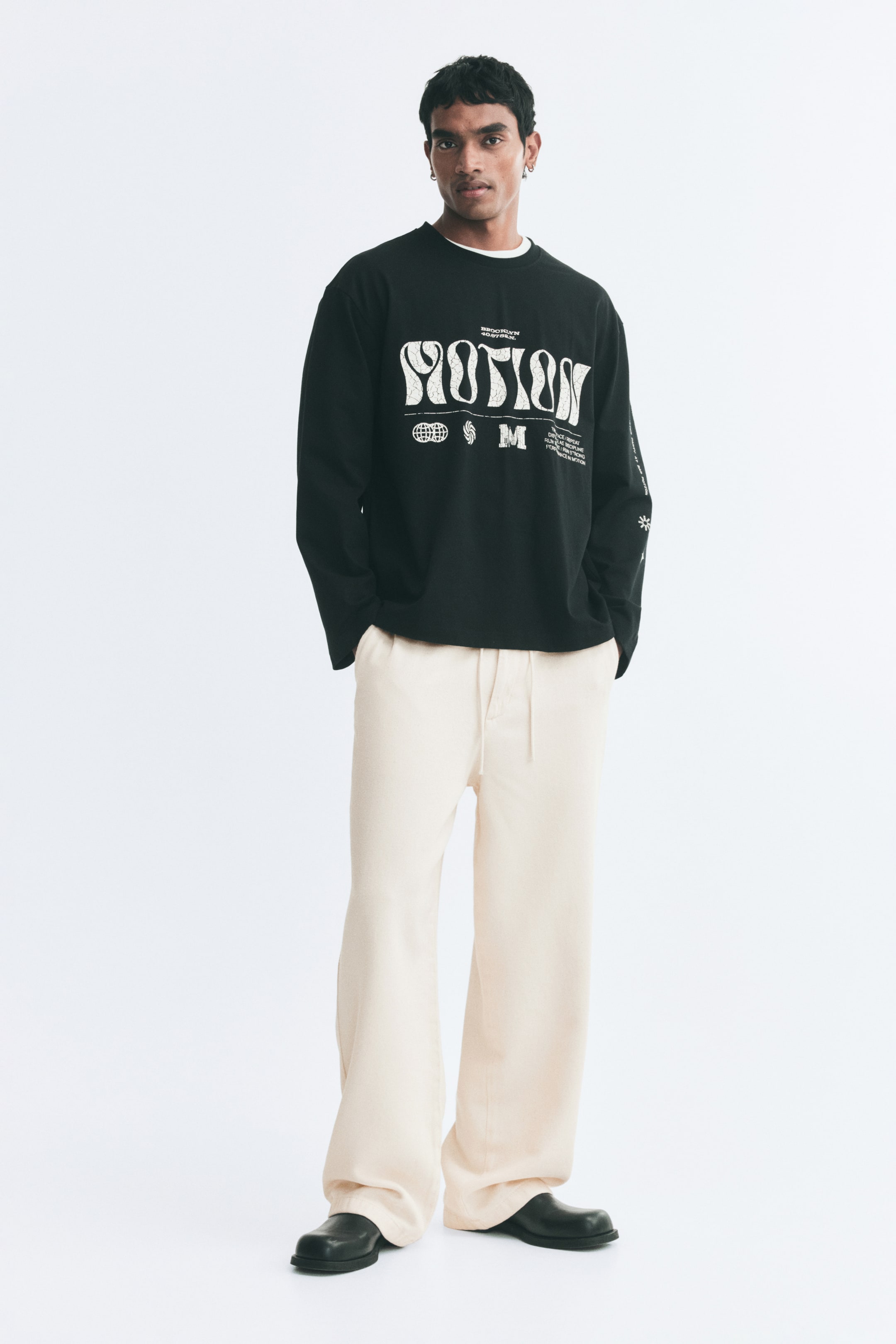 View larger image: Relaxed-fit pants in an off-white shade, featuring an elasticated drawstring waist and diagonal side pockets, are being worn by the model. The model is standing with hands in the pockets, styling the pants with a black graphic long-sleeve T-shirt and black shoes.