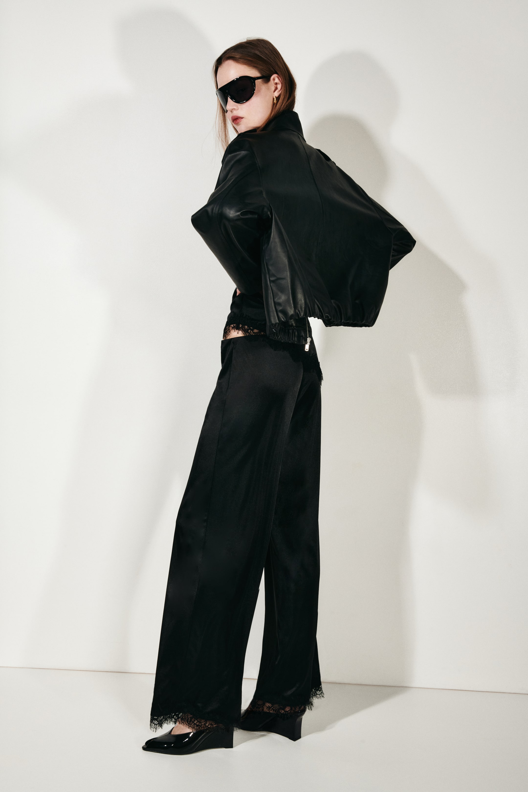 Agrandir l'image: Une veste bomber noire raccourcie, avec des manches volumineuses et un ourlet élastiqué, est portée sur un pantalon large en satin noir. Le pantalon présente une bordure en dentelle noire à la taille et à l'ourlet et est associé à des chaussures compensées à bout pointu noires, l'ensemble étant présenté sous un angle de côté-arrière.