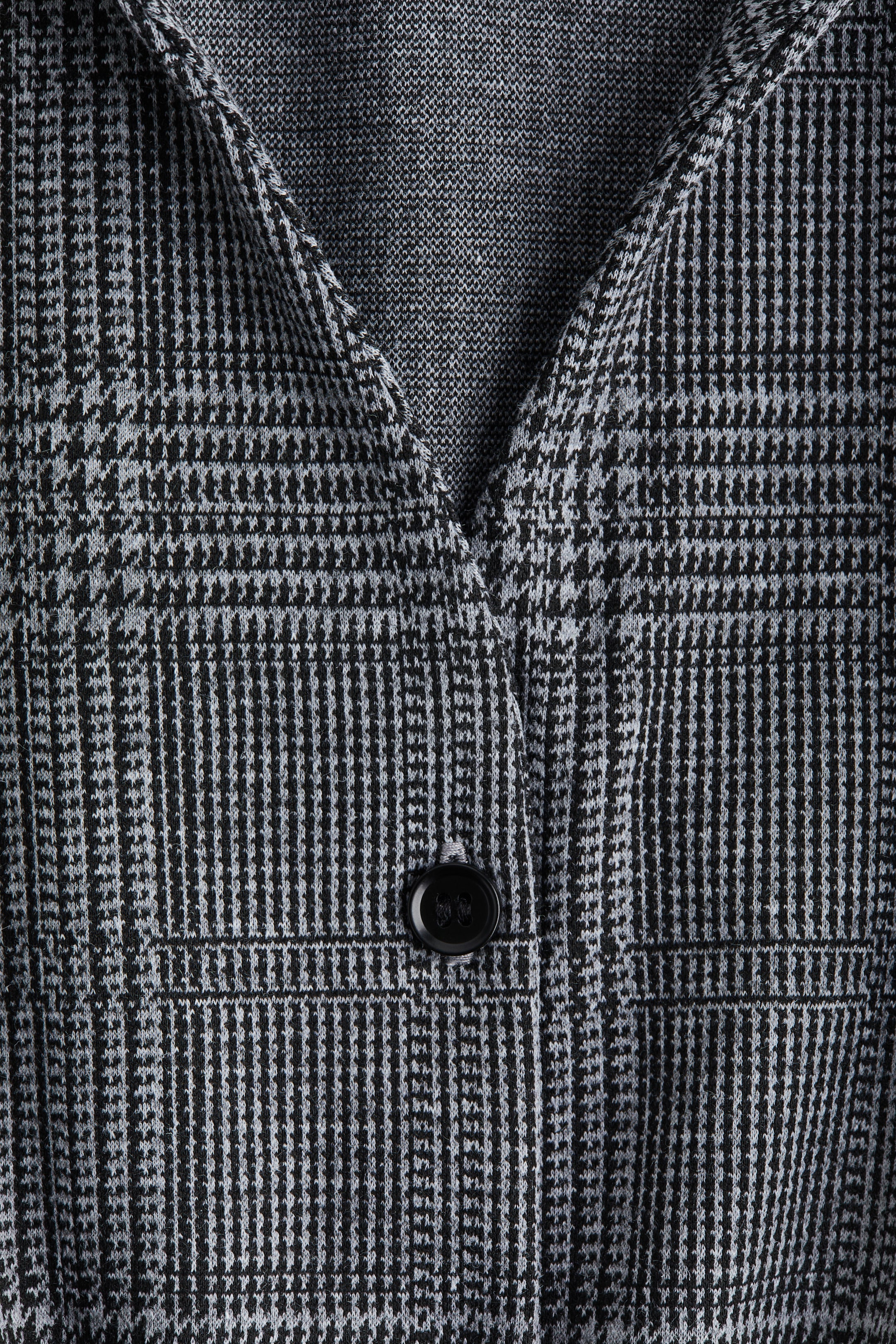 View larger image: A front view of a garment is presenting a black and light grey glen plaid knit pattern, and is designed with a V-shaped neckline and a single black button closure.