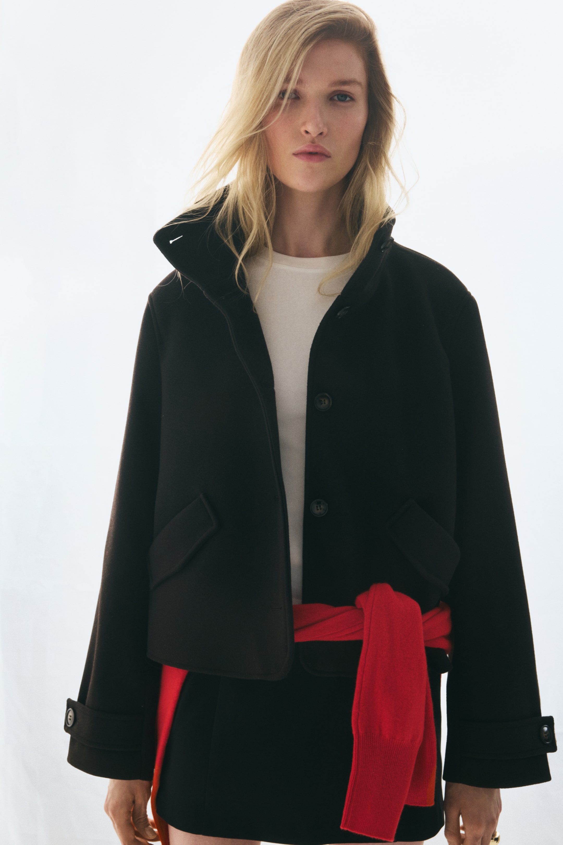 View larger image: A black short jacket in woven fabric is shown from the front, featuring a high funnel collar, a button-front closure, and diagonal front pockets with flaps. The jacket is worn open over a white crew-neck top with a red sweater tied around the waist, highlighting its boxy silhouette and button-tab cuffs.