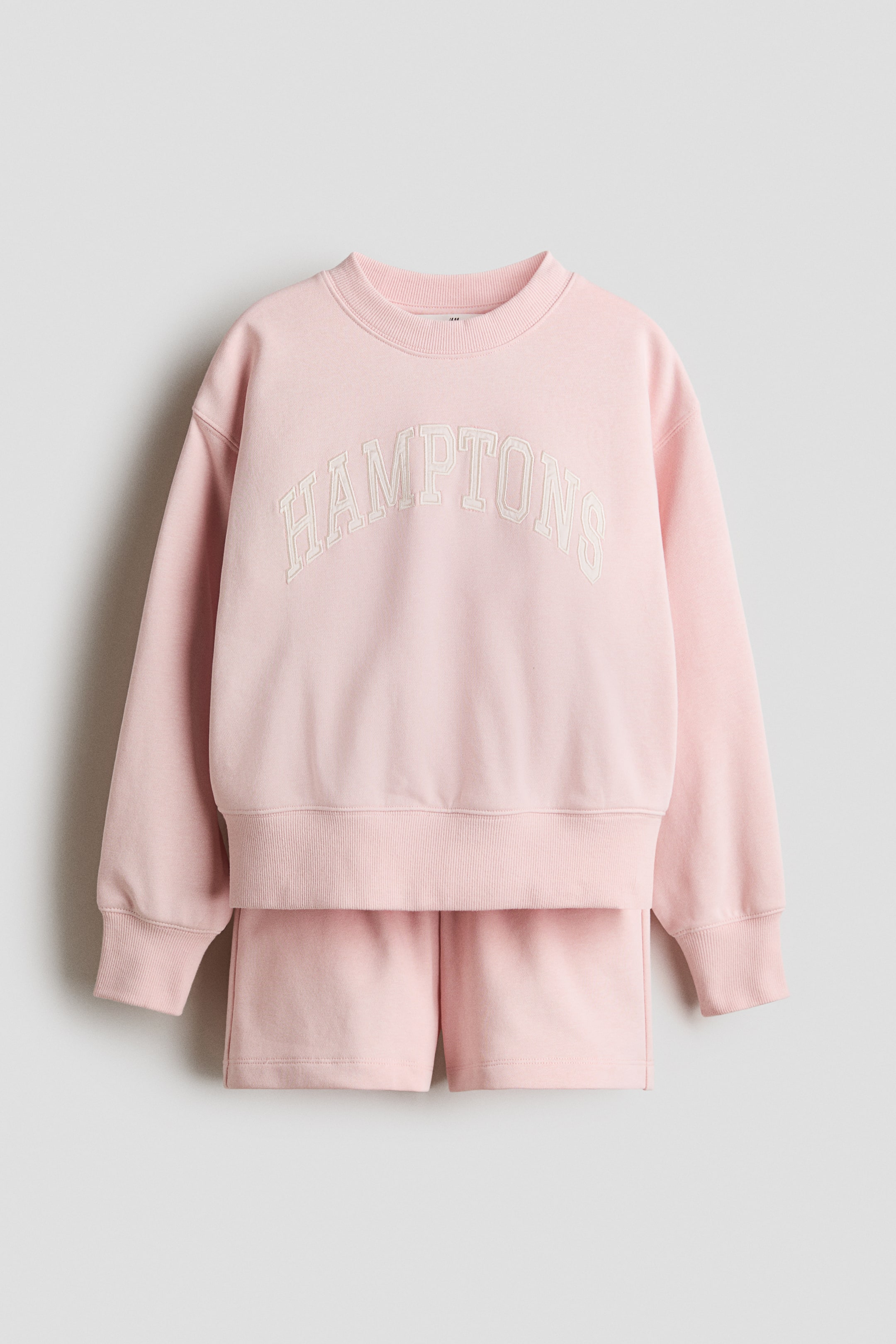 Grösseres Bild ansehen: Ein rosa langärmliges Sweatshirt und ein dazu passendes Shorts-Set werden von vorne präsentiert. Das Sweatshirt verfügt über einen gerippten Rundhalsausschnitt, überschnittene Schultern, gerippte Bündchen und einen gerippten Saum, mit "HAMPTONS" in Weiß auf der Brust gedruckt. Die passenden Shorts haben einen elastischen Bund.