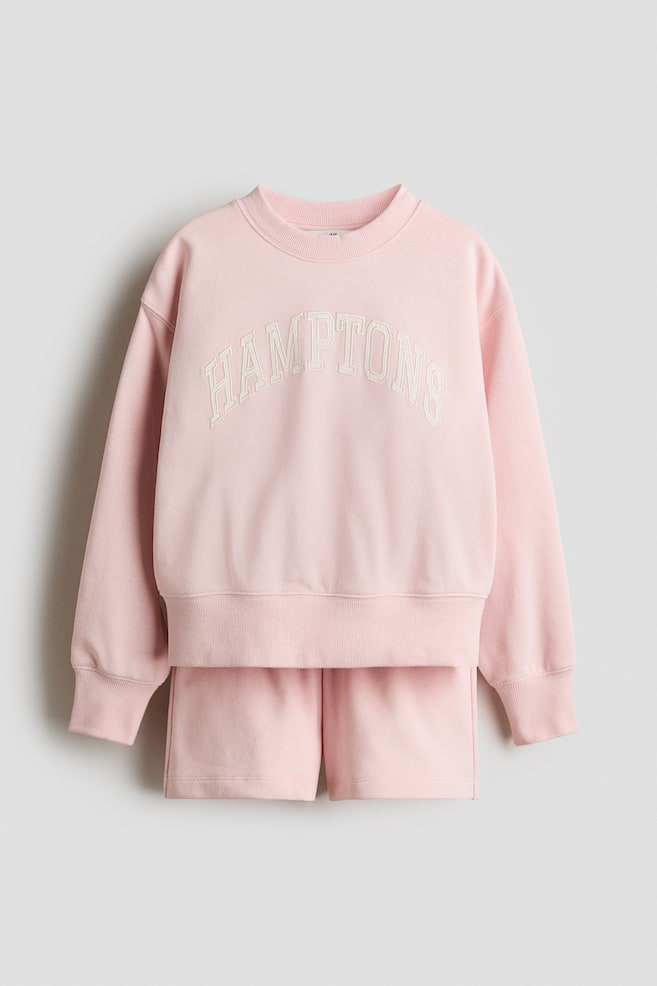 A pink long-sleeved sweatshirt and matching shorts set is shown from the front. The sweatshirt features a ribbed crew neckline, dropped shoulders, ribbed cuffs, and a ribbed hem, with "HAMPTONS" printed in white across the chest. The coordinating shorts present an elasticated waist. - 1