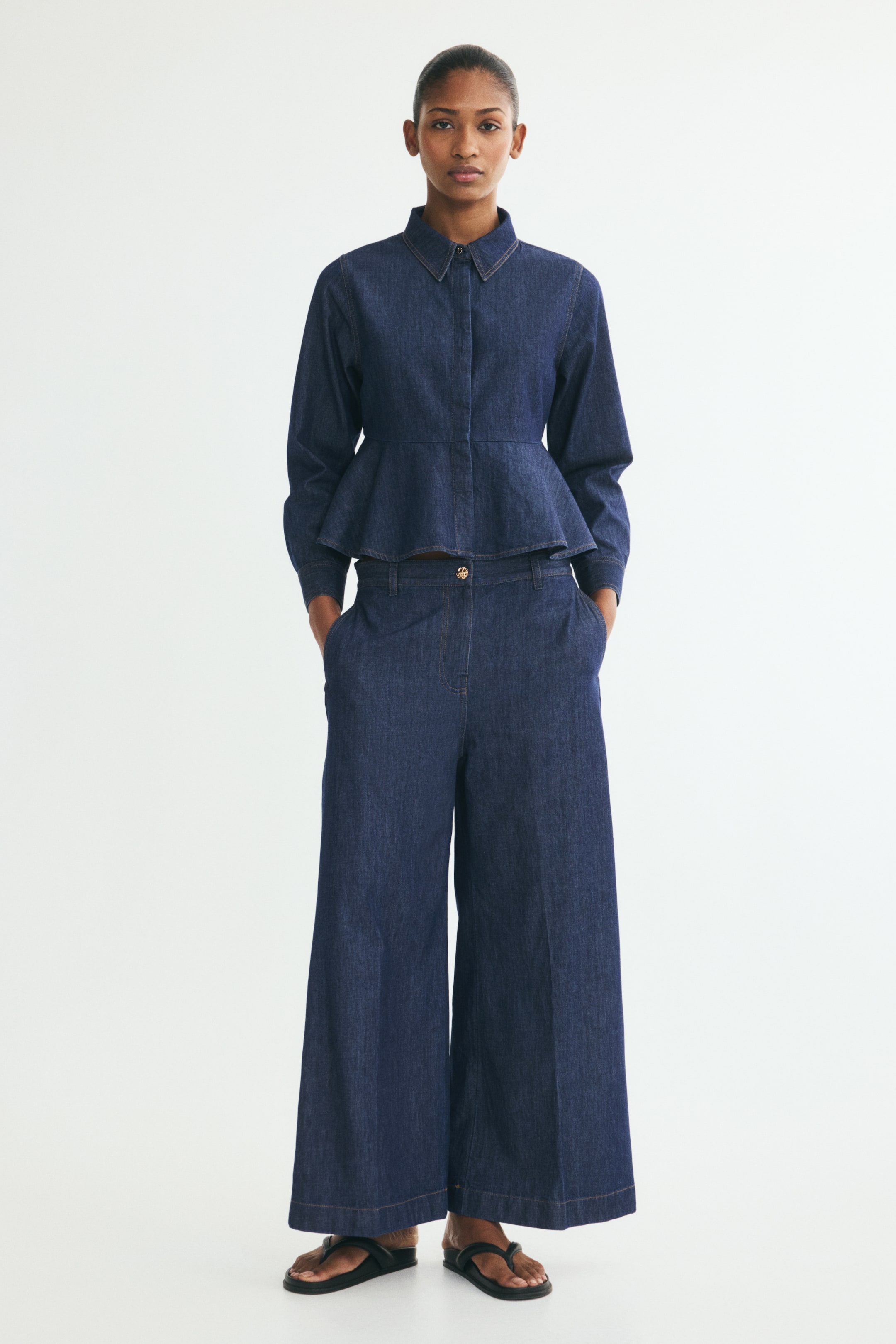 View larger image: These dark blue, wide-leg denim jeans are shown from the front, featuring a regular waist, a zip fly with a shiny, irregular-shaped gold button, and subtle creases running down the centre of each leg. The loose-fitting jeans are styled with a matching dark blue denim peplum shirt, emphasising their wide, structured silhouette that drapes to the feet.