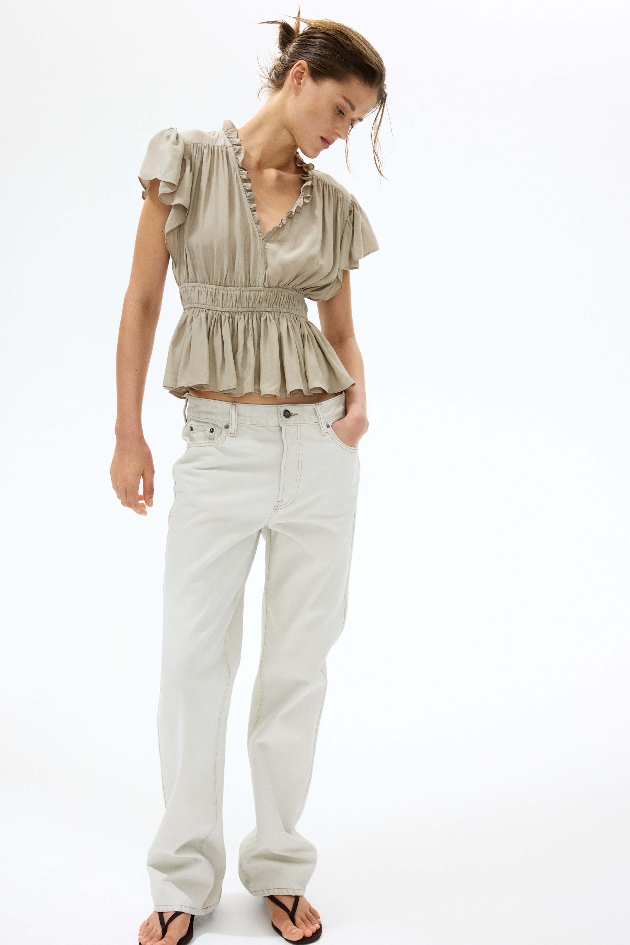 Se større billede: En beige peplumtop fremviser en flæsekrave i V-hals, flæseærmer med kapærmer og en smock-elastisk talje, der skaber en udsvingende silhuet. Den styles med off-white jeans med lige ben, som er kendetegnet ved fremtrædende kontrastsyninger.