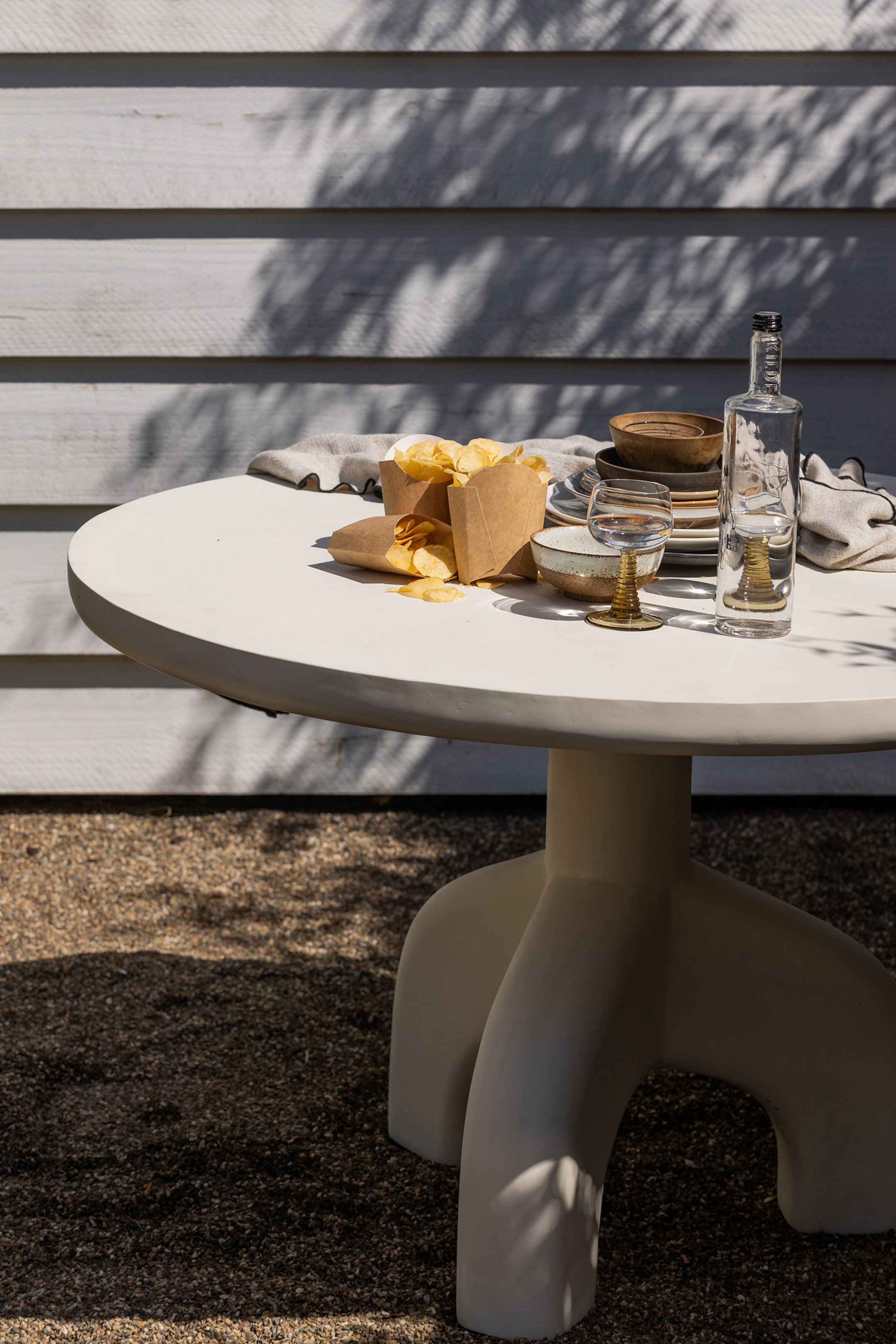 Agrandir l'image: Une table ronde de couleur claire est vue d'un angle légèrement surélevé, mettant en valeur son plateau lisse et un pied central qui se ramifie en trois pieds épais et arrondis. Divers plats, verres, une bouteille et des sachets de snacks reposent sur son plateau.