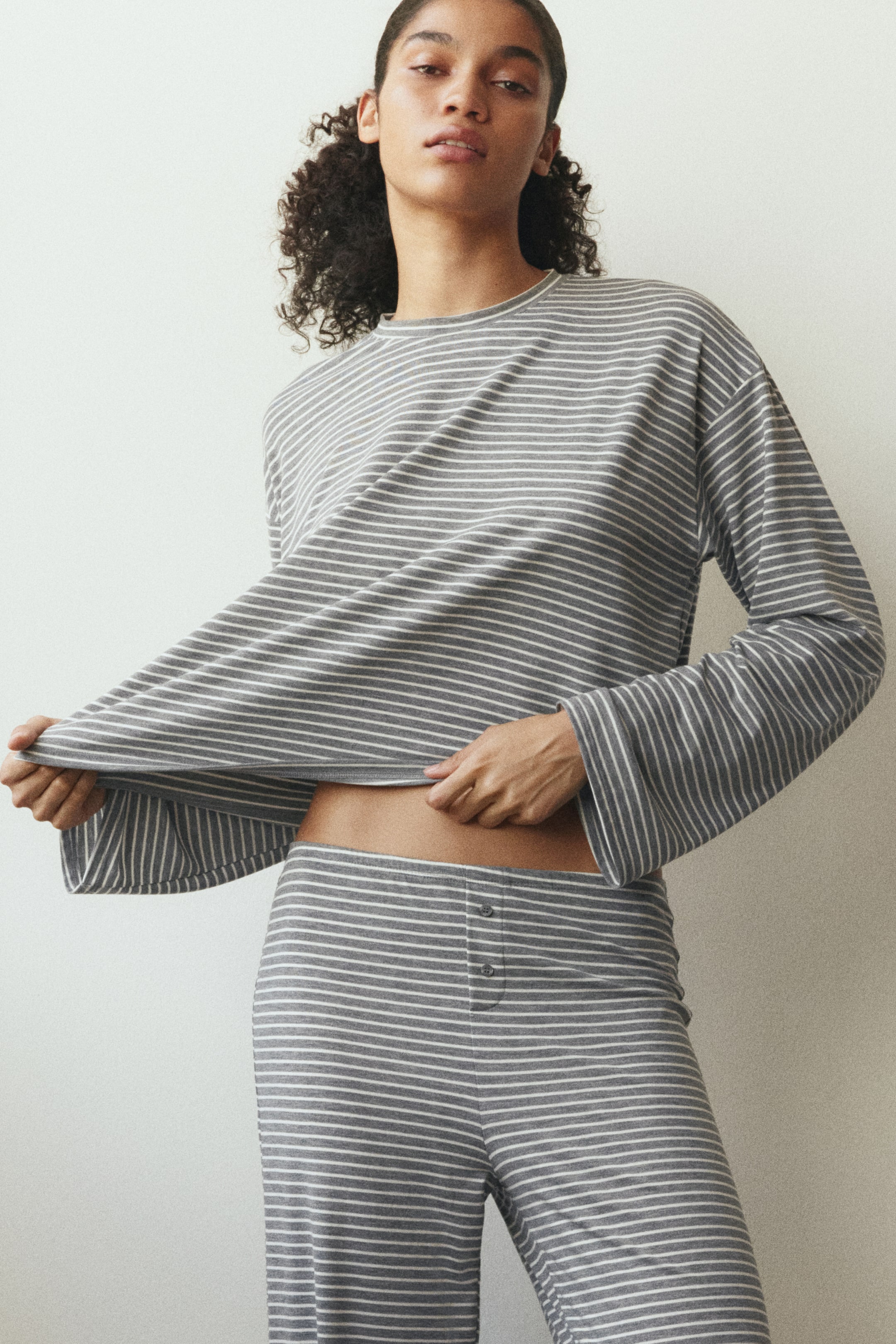 Se større billede: Et gråt og hvidt stribet loungewear-sæt, bestående af en langærmet top med rund hals og lige benede bukser med en knaplukning med tre knapper, er i brug. Modellen trækker topmens kanten op med begge hænder, så hendes mave samt buksernes taljedetalje kommer til syne.