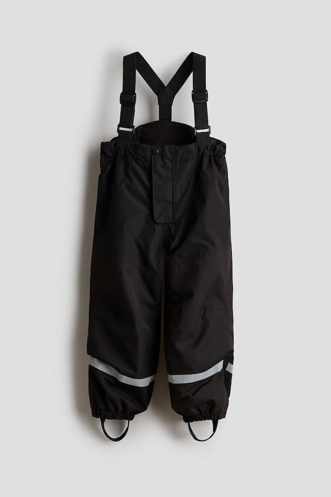 A pair of black children's snow pants is facing front, featuring adjustable elastic suspenders and an elasticated waistband with a snap-button placket. The pants are showing horizontal reflective bands near the lower legs and elasticated ankle cuffs with underfoot stirrup straps. - 1
