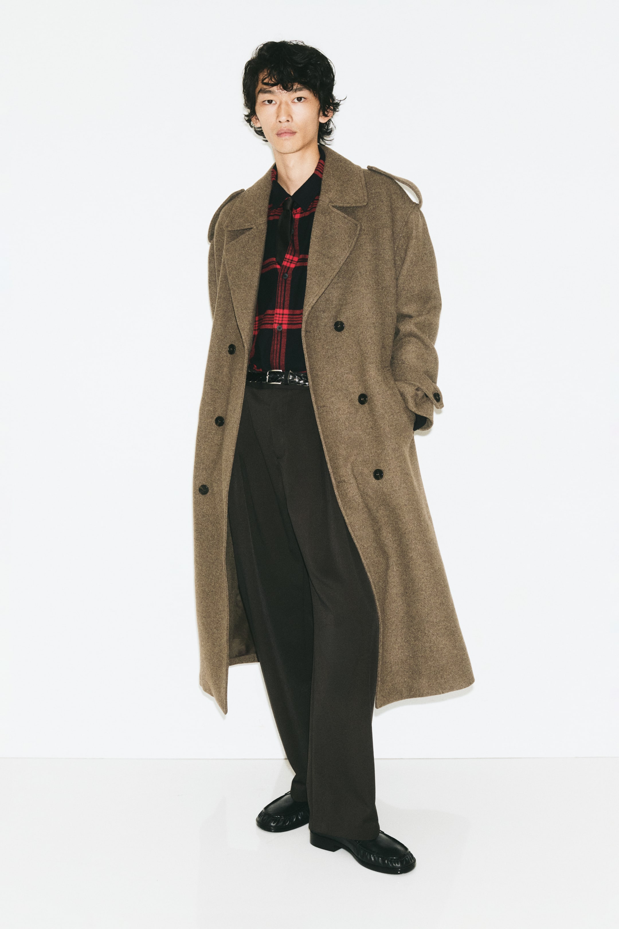 Größeres Bild anzeigen: Locker geschnittene, maßgeschneiderte Hosen aus dunkelbraunem Twill werden vom Model getragen und zeichnen sich durch vordere Bundfalten sowie eine entspannte Barrel-Leg-Silhouette aus. Die Hose ist mit einem in den Bund gesteckten rot-schwarzen Karohemd, einem schwarzen Ledergürtel, einem langen braunen zweireihigen Mantel und schwarzen Lederschuhen kombiniert.