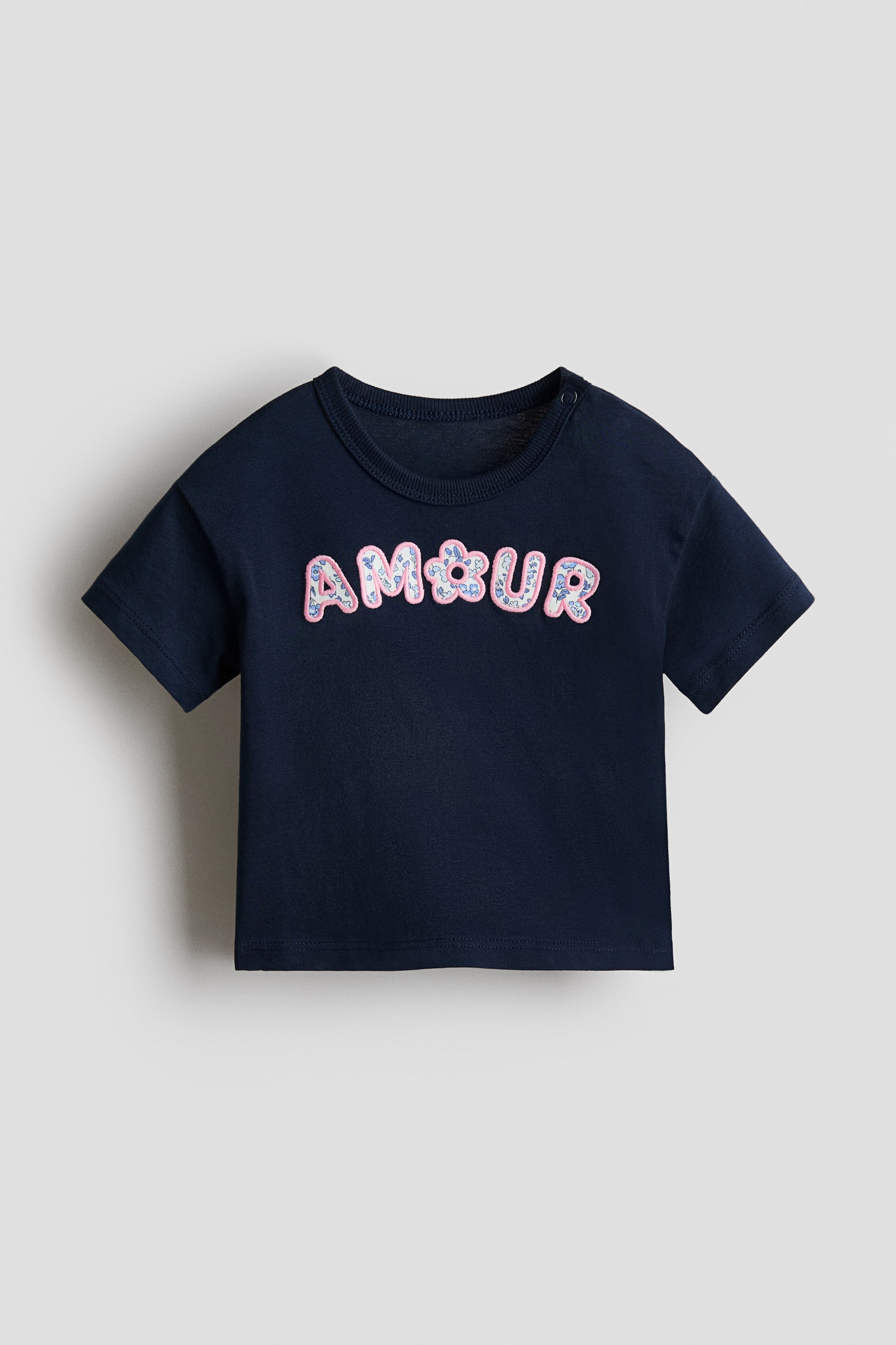 View larger image: A navy blue T-shirt is front-facing, featuring short sleeves, a crew neck, and a snap button on the left shoulder. It displays "AMOUR" on the chest, with the letters outlined in pink and filled with a light blue and pink floral pattern.