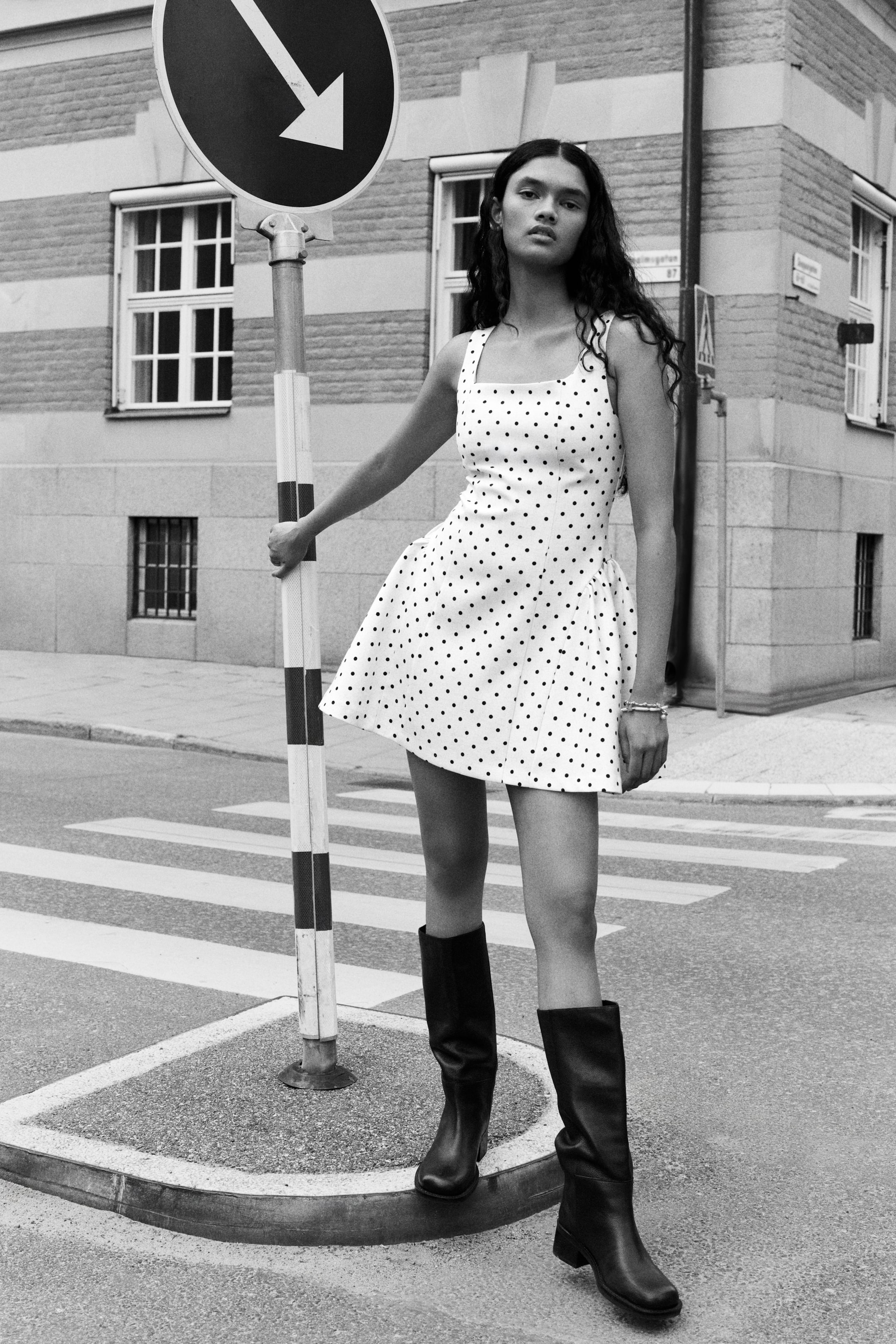 View larger image: A short, polka-dot patterned dress featuring a square neckline, wide shoulder straps, a fitted bodice, and a flared skirt with gathered panels at the hips is being worn by the model. The garment is styled with knee-high black boots.
