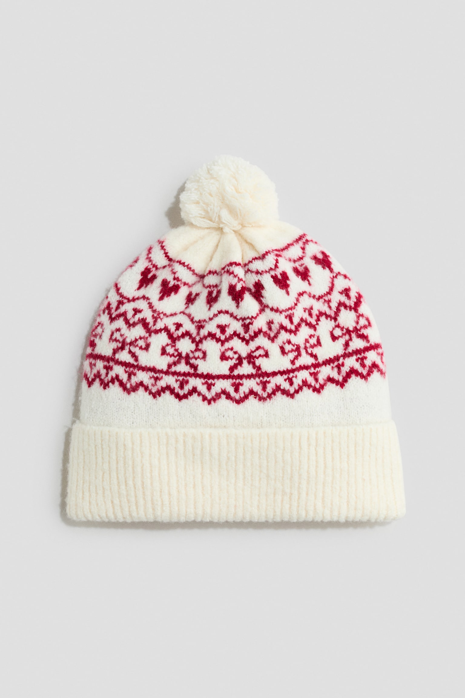 En stickad mössa är vänd framåt och visar en benvit bas med ett rött Fair Isle-mönster av rosetter och geometriska motiv. Mössan har en ribbstickad uppvikt mudd nedtill och en matchande krämfärgad stickad tofs upptill.