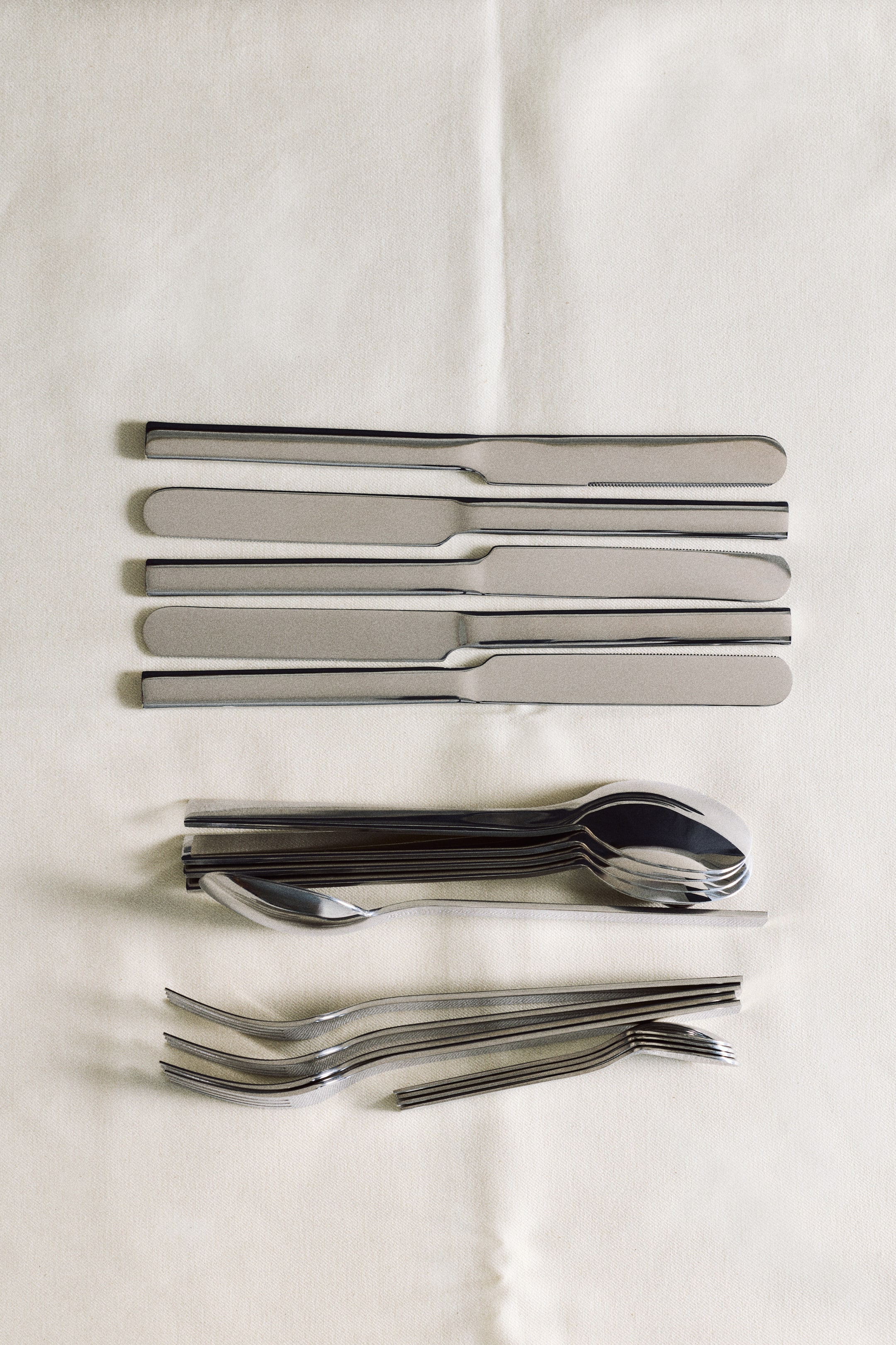 Vis større bilde: Dette bestikksettet består av elegante deler i rustfritt stål med flate håndtak, arrangert i ryddige rader sett ovenfra. Fem kniver ligger horisontalt i vekslende retninger over stablede grupper av skjeer og gafler i ulike størrelser.