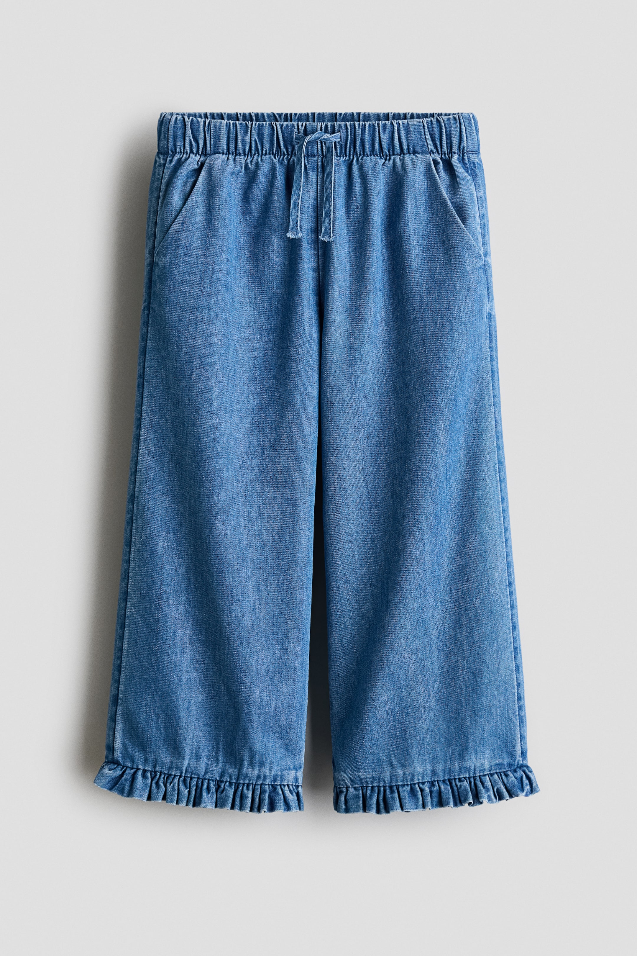 View larger image: Blue denim wide-leg pants feature an elasticated, ruched waistband with a central drawstring tie at the front. They are designed with two diagonal slash pockets and ruffled hems at the cropped leg openings.