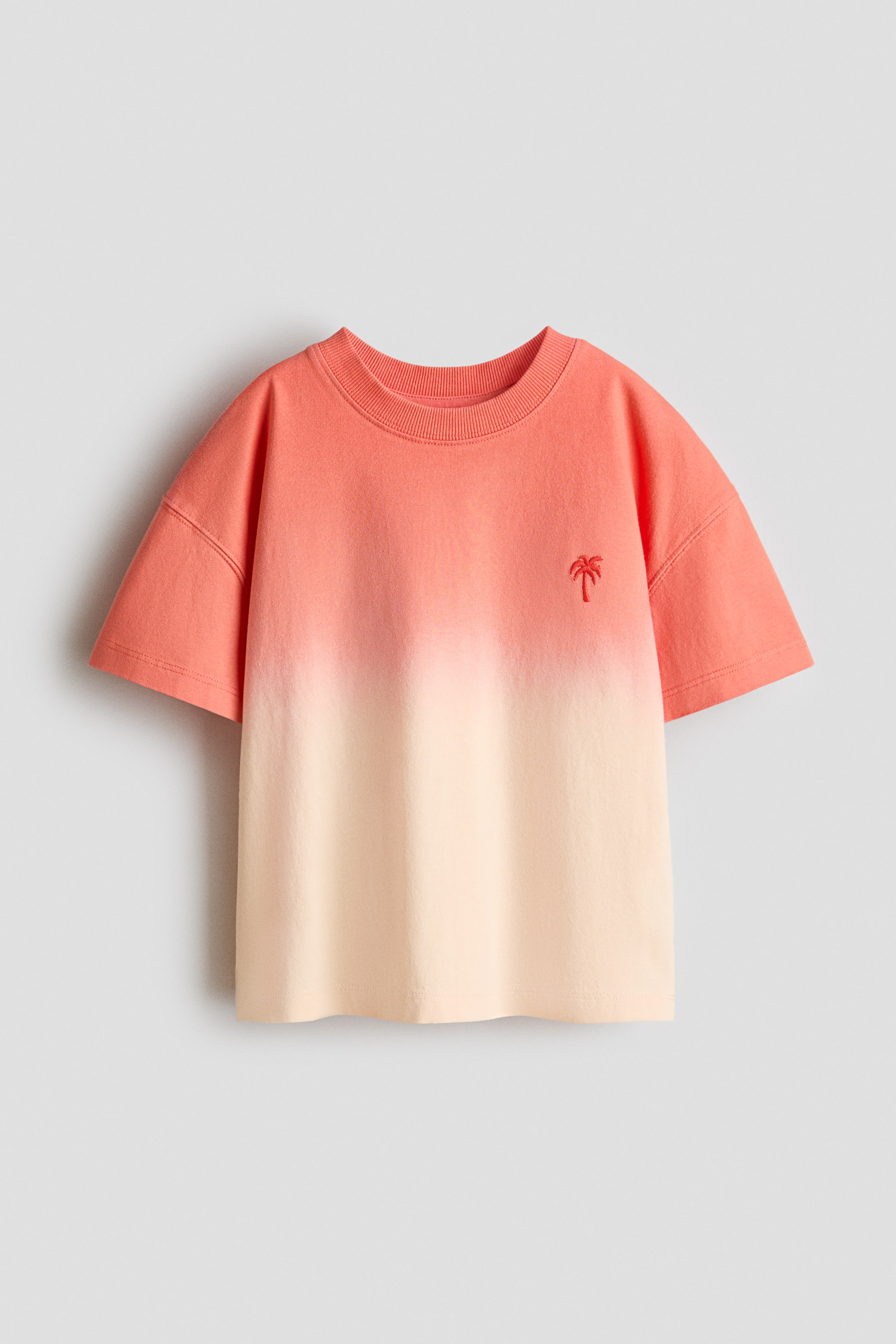 View larger image: An oversized T-shirt, viewed from the front, is displaying an ombré design transitioning from coral at the top to light cream at the bottom. It features a ribbed neckline, dropped shoulders, a straight-cut hem, and a small red palm tree motif embroidered on the left chest.