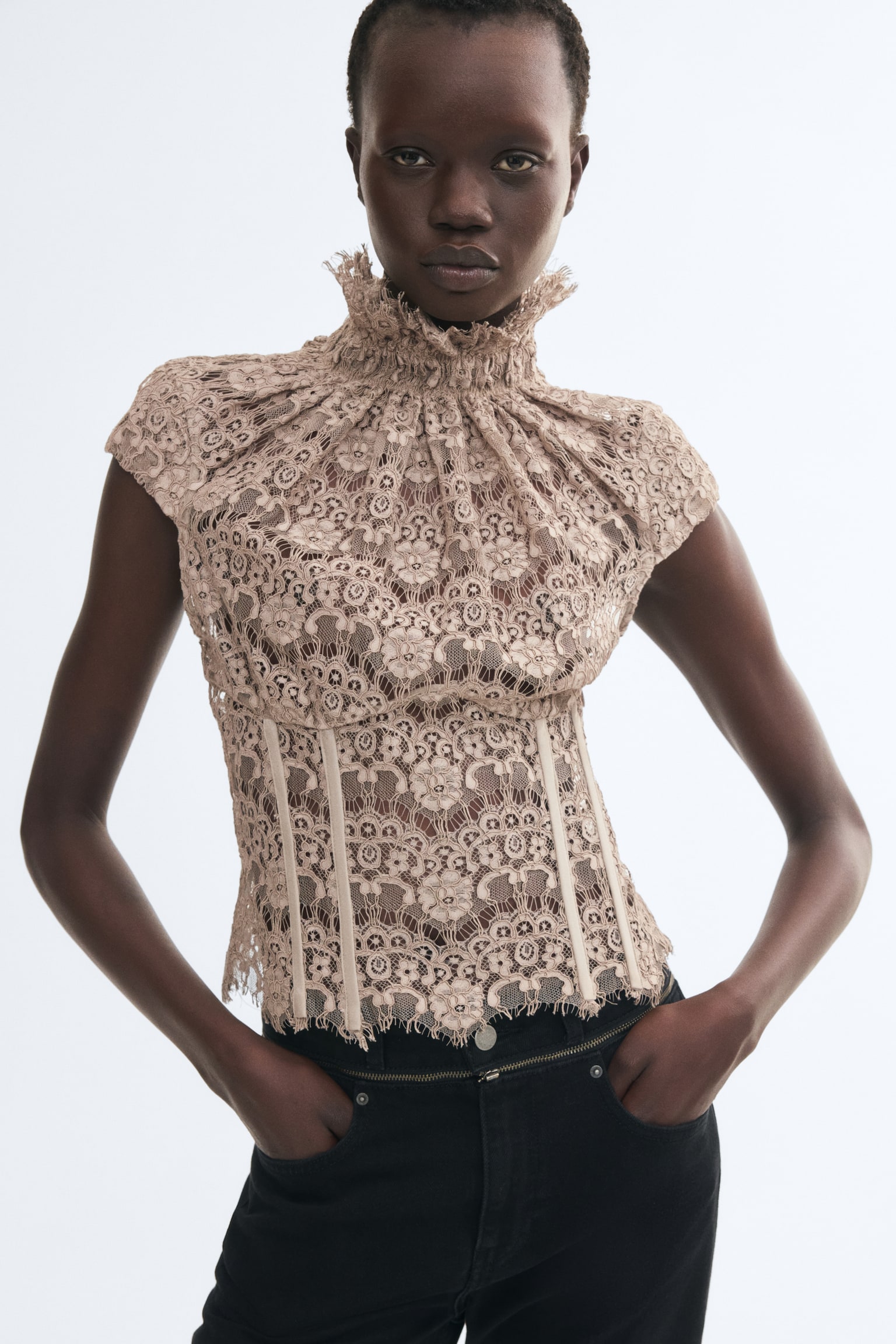 A beige lace corset top is being worn by the model, featuring a high, gathered neckline with a frill trim, cap sleeves, and visible vertical boning along the front waist. The top is styled with black jeans, with the model placing their hands in the front pockets. - 1