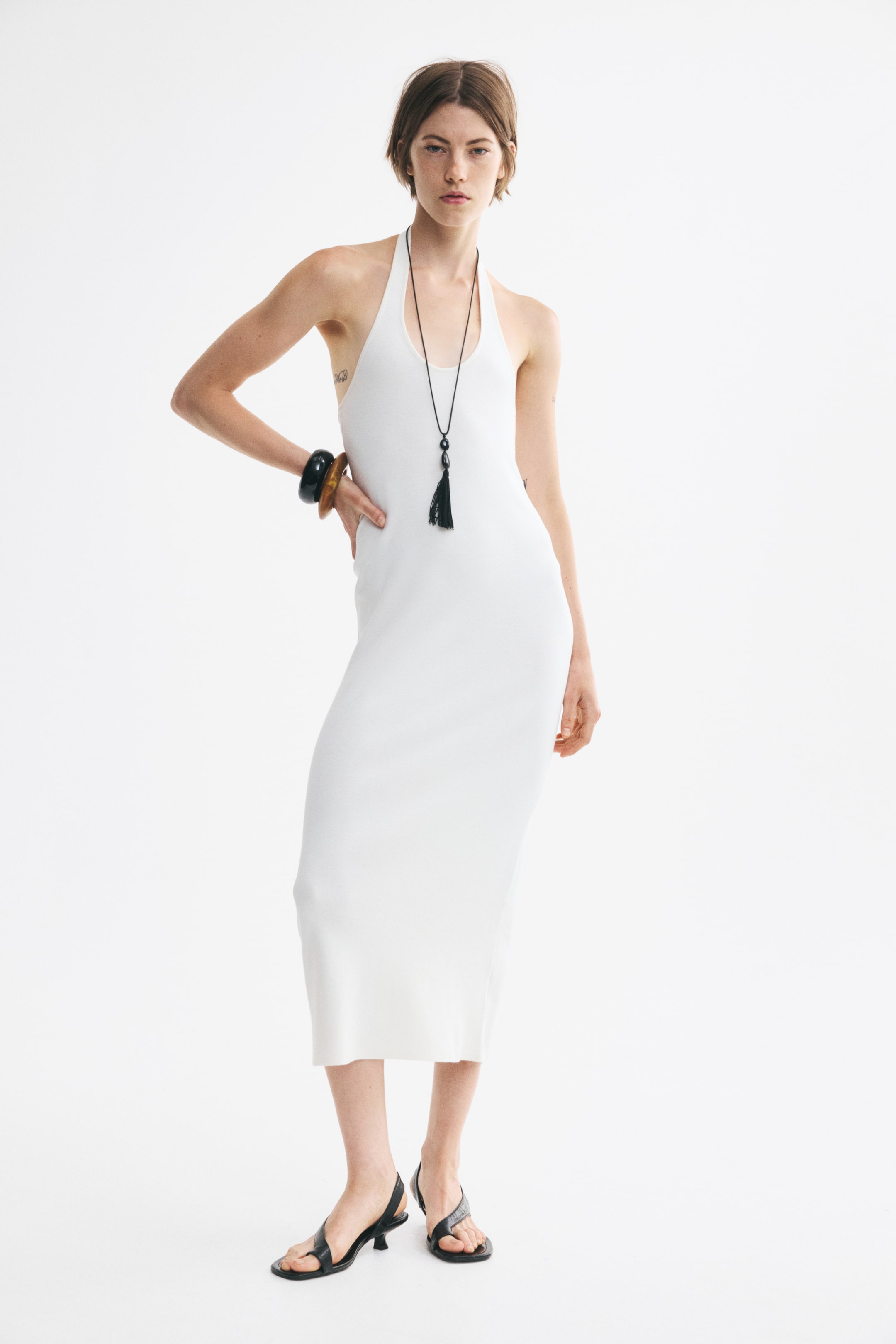 View larger image: A white ribbed-knit midi dress features a halter neck strap and a scoop neckline, viewed from the front. The sleeveless dress clings to the body in a form-fitting silhouette and drapes to a straight, mid-calf hem.