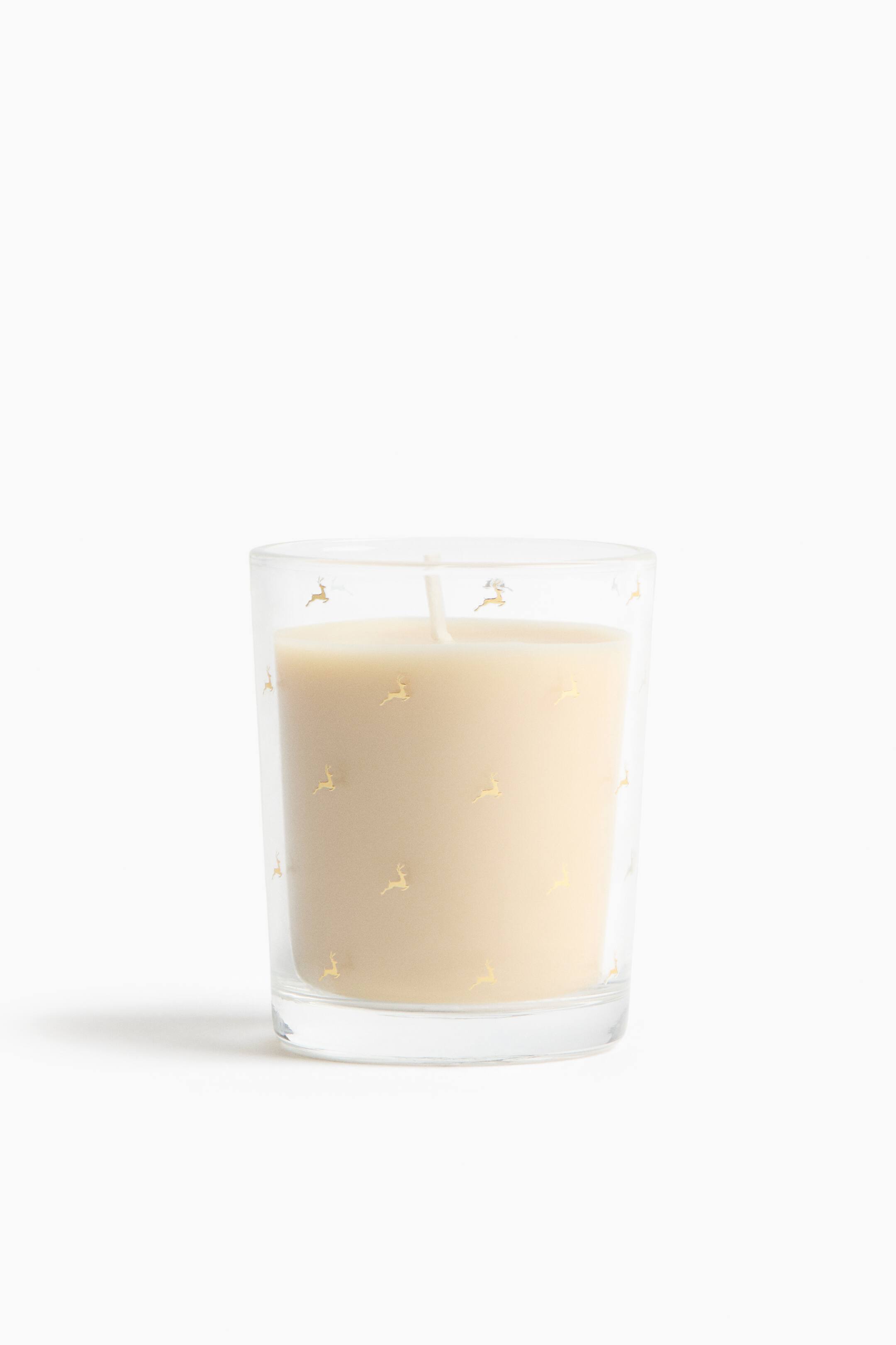 View larger image: A cream-coloured wax candle is placed inside a clear, slightly tapered glass tumbler featuring small, scattered gold reindeer motifs. The product is shown front-facing, with a single centred white wick rising from the smooth wax.