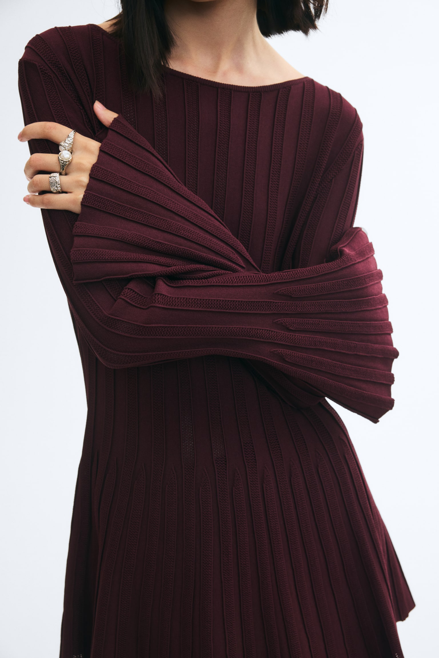 Silver-toned metal rings in textured, ornate designs, including one featuring a pearly white bead, are worn on the modelâs fingers. The rings are styled alongside a long-sleeved, ribbed burgundy knit garment with pleated sleeves. - 1