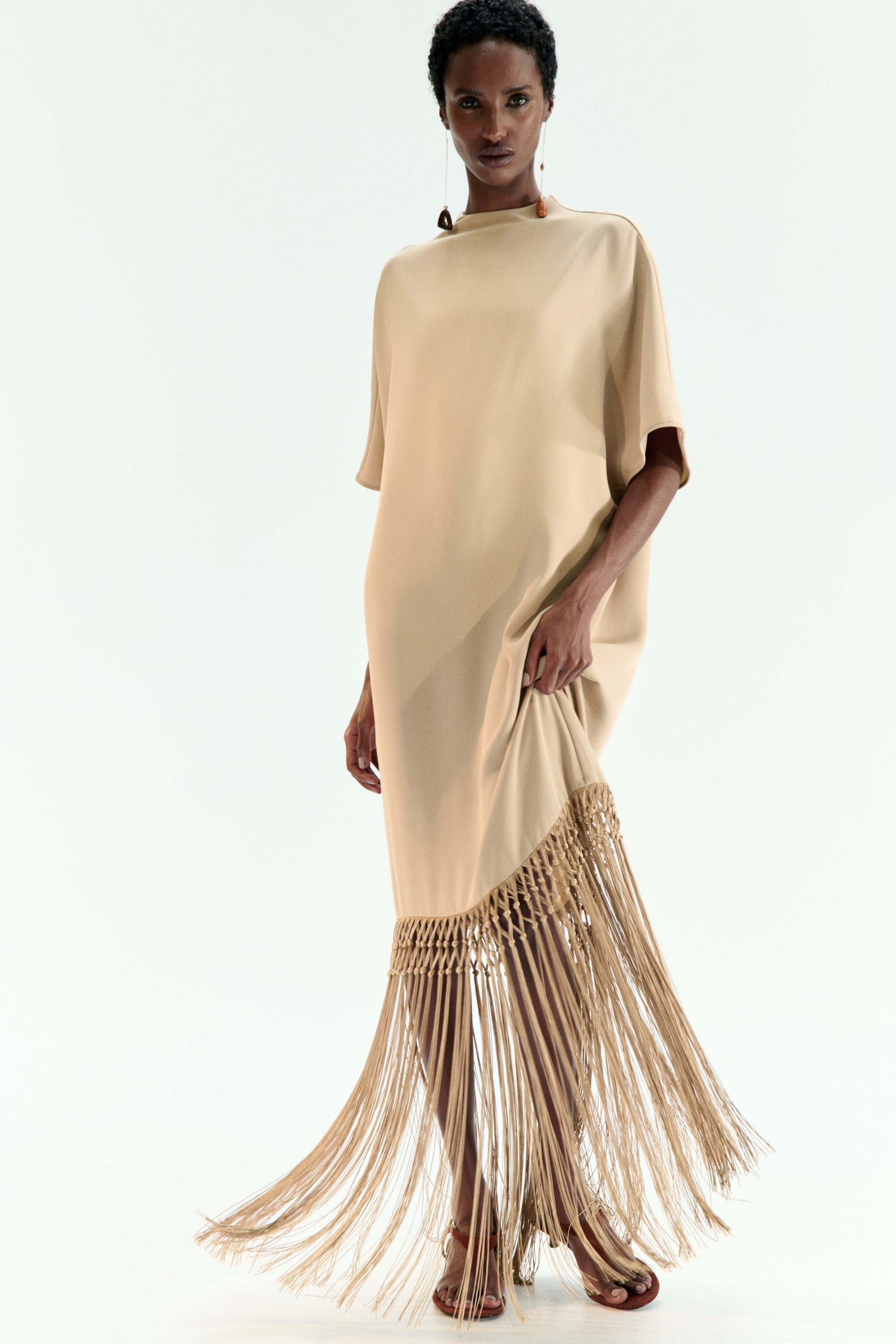 View larger image: A beige loose-fitting maxi dress is presented from a front-side angle, featuring a high neckline and wide, short sleeves. Its asymmetrical hemline is adorned with a thick, woven macrame border, from which long, flowing fringe strands are extending.
