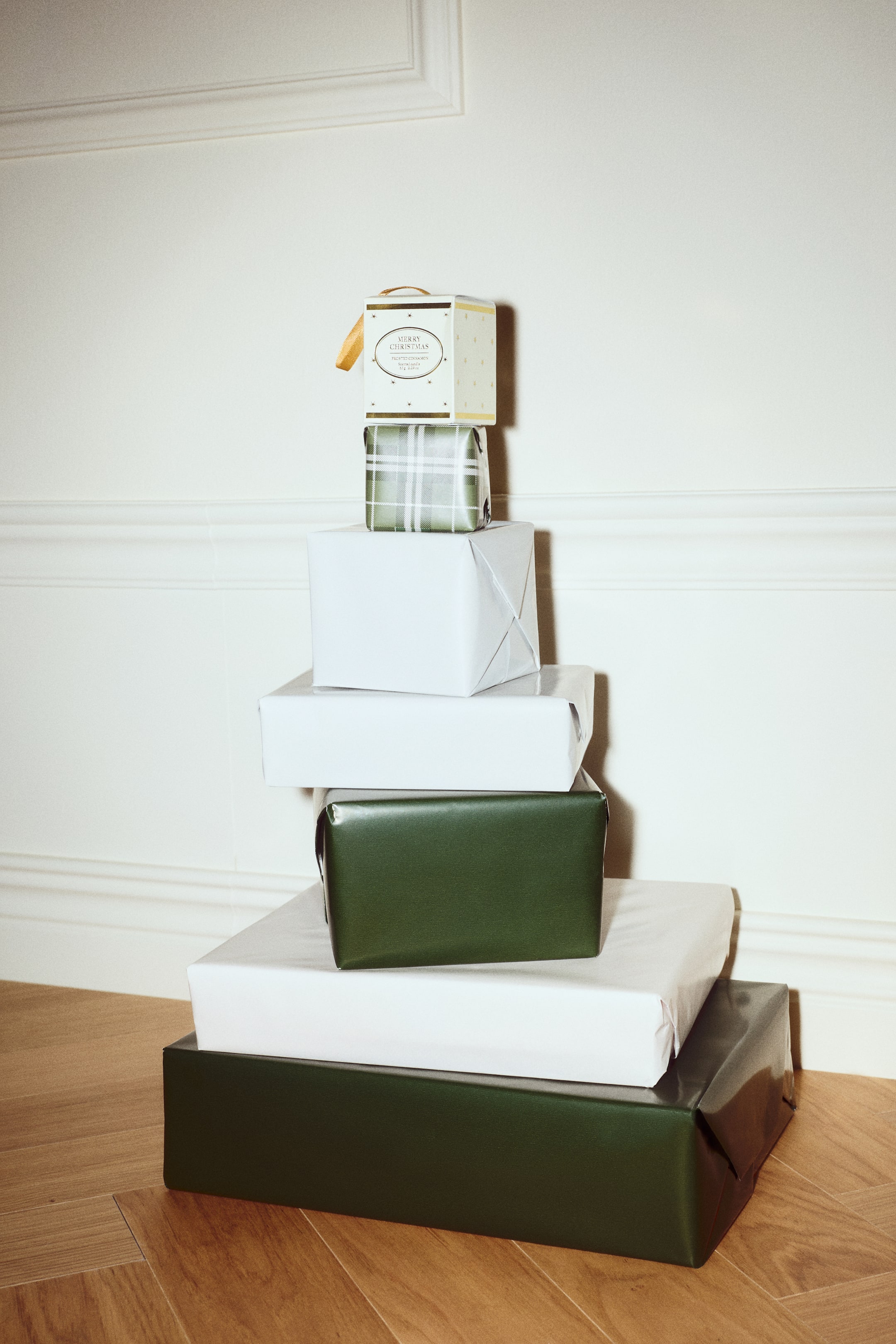 Agrandir l'image: Une élégante boîte blanche en forme de cube contenant une bougie parfumée repose au sommet d’une pile de cadeaux emballés, orientée vers l’avant. La boîte présente de délicats motifs d’étoiles dorées, une étiquette circulaire sur la face avant et une boucle de ruban doré suspendue en haut.